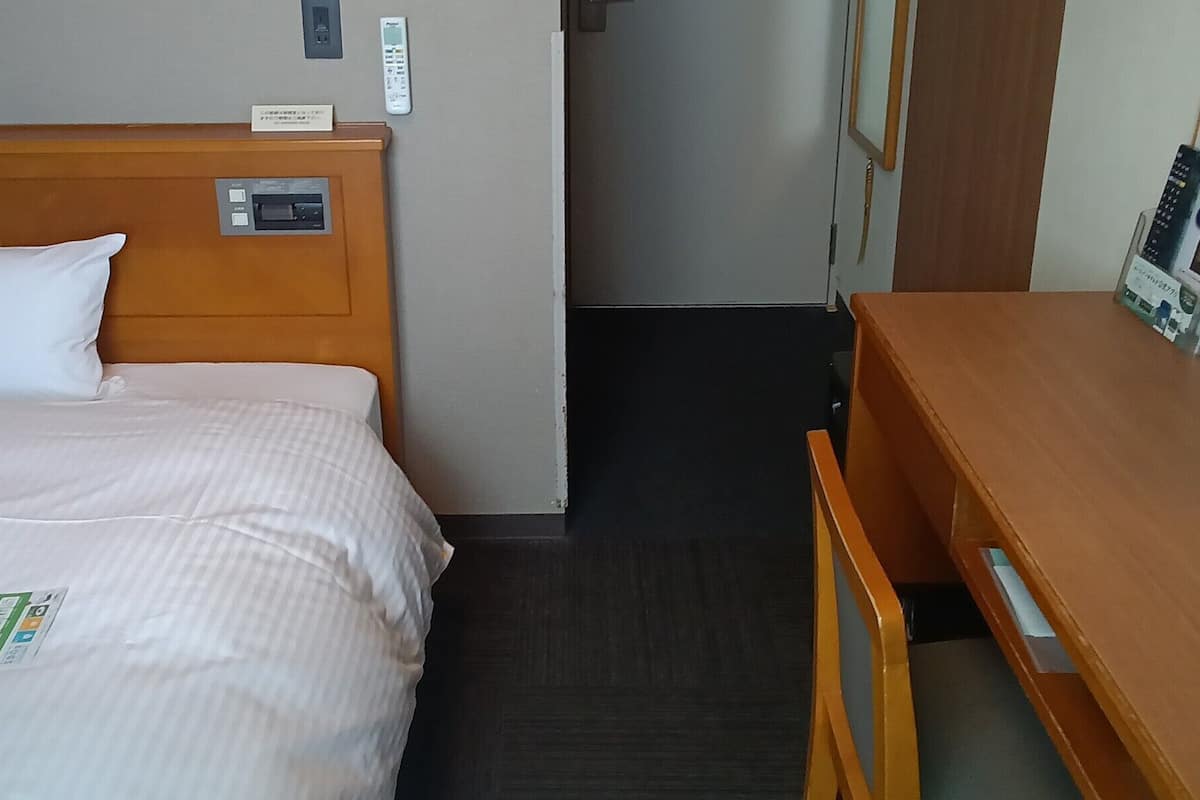 A hotel room with a bed, a wooden nightstand, a desk, a chair, and a door.