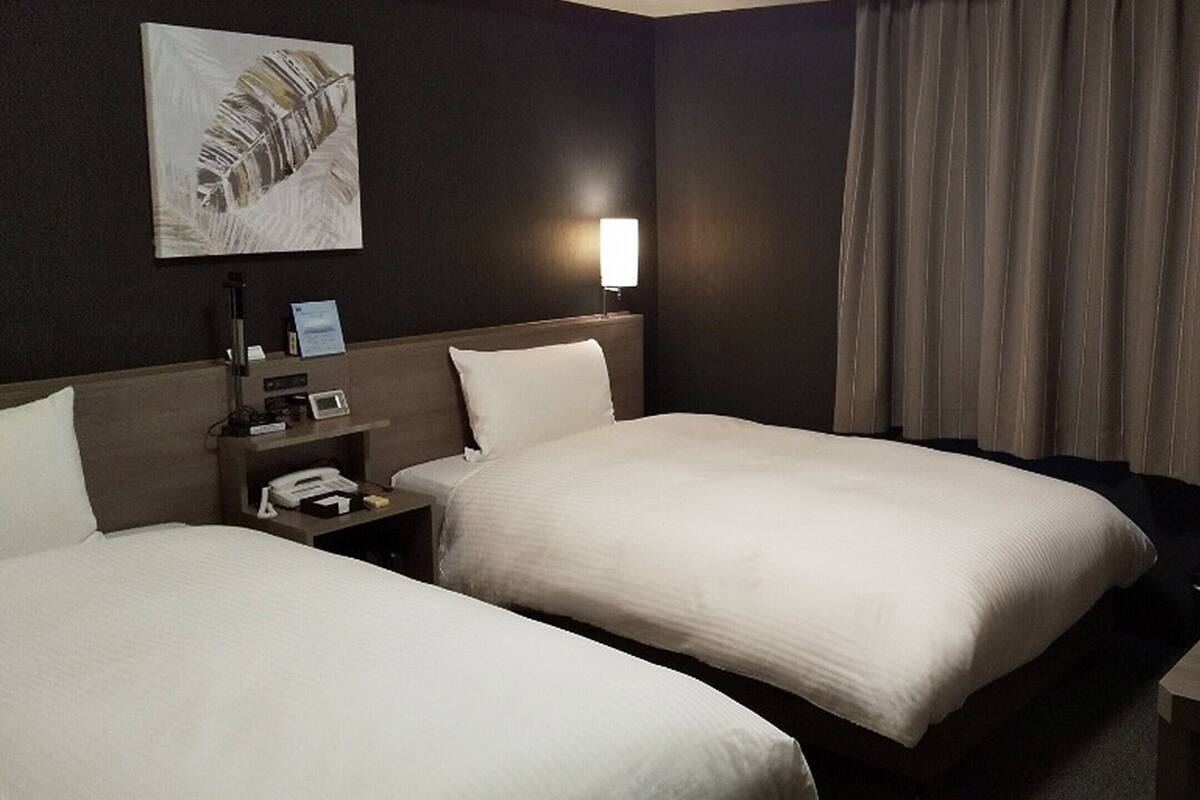 Camera d'albergo con due letti, un comodino con una lampada, un quadro appeso e una finestra con tende.