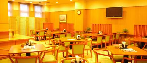 A hotel dining area with multiple tables and chairs, a clock on the wall, and a television mounted on the wall.