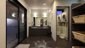 Un baño moderno con suelo de madera oscura, un espejo grande, lavamanos dobles, un reloj y una zona de lavandería con cestas.