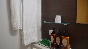 A bathroom with a white towel hanging on a rack, a glass shelf with a white cup, and three bottles of body soap and shampoo.