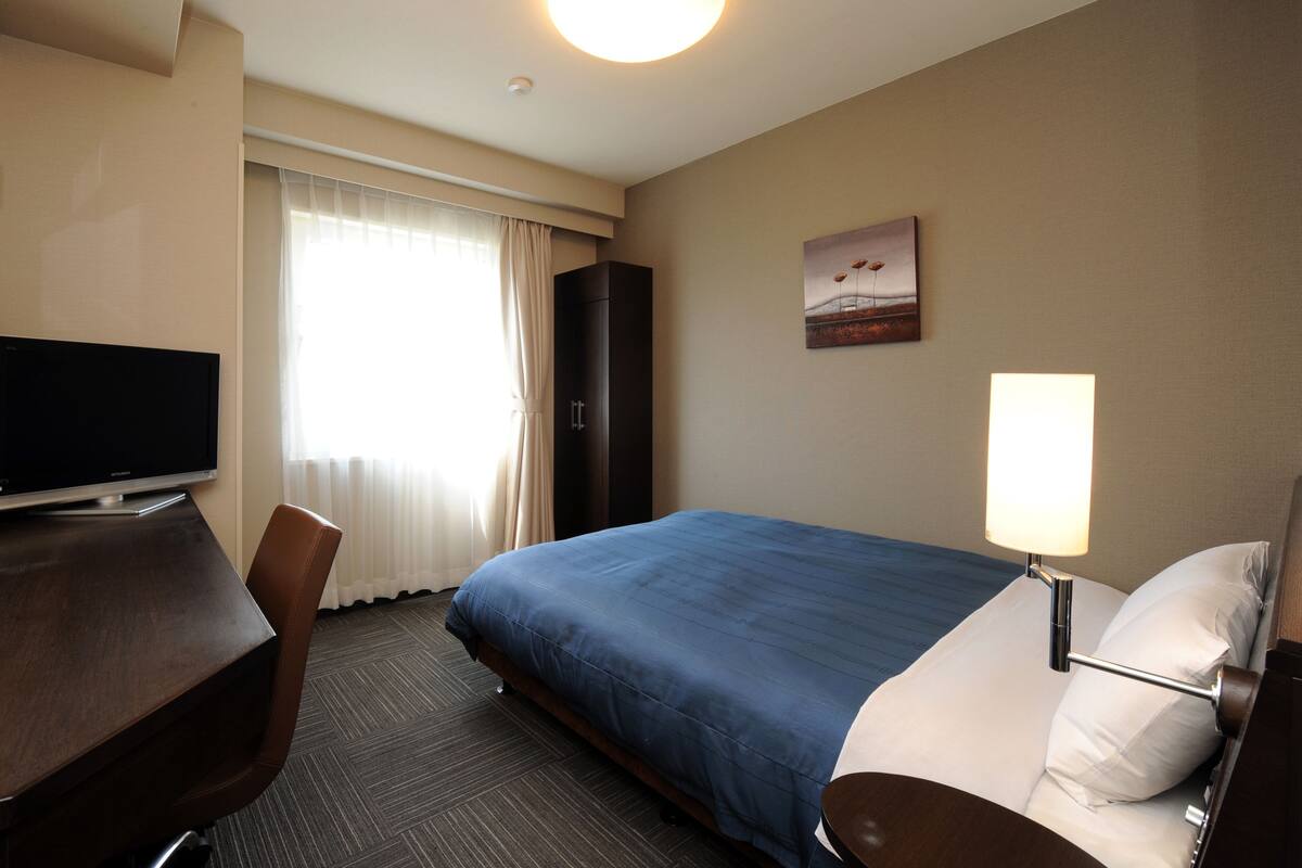 A hotel room with a bed, a desk, a chair, a television, a lamp, and a painting on the wall.