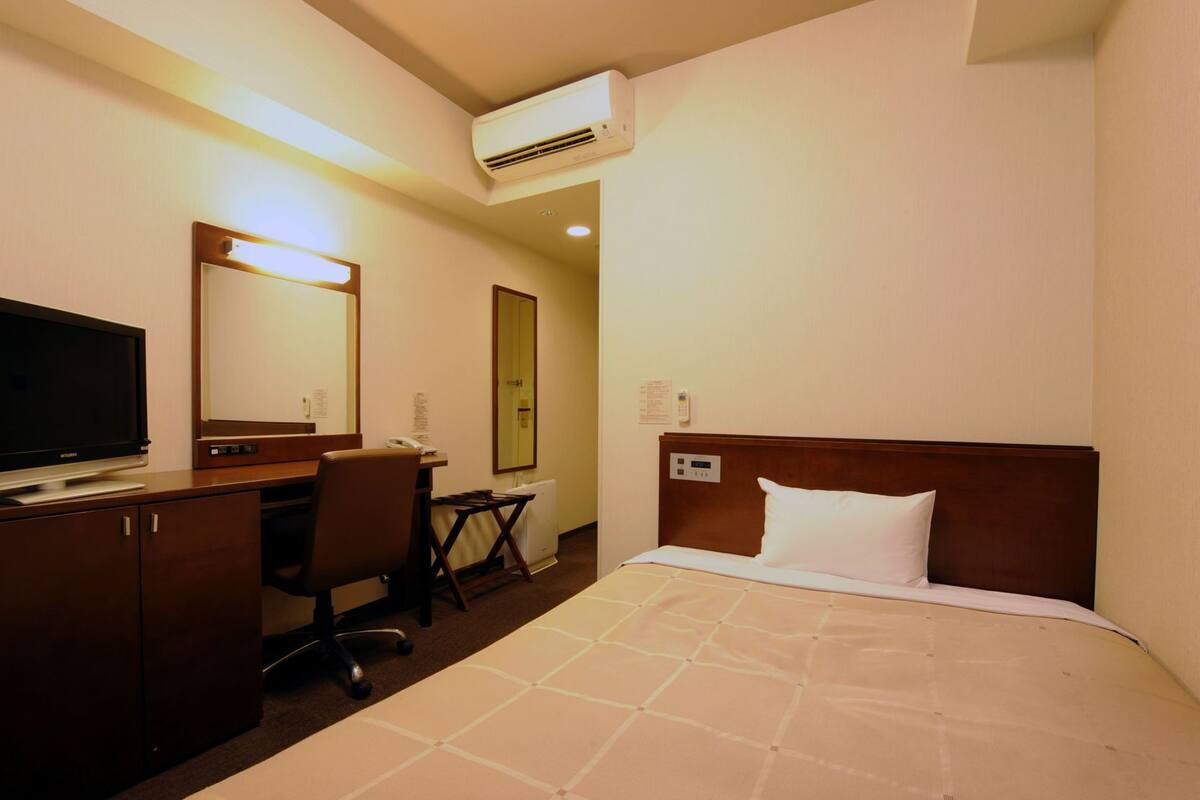 A hotel room with a bed, a desk with a television, a chair, an air conditioning unit, and a mirror.