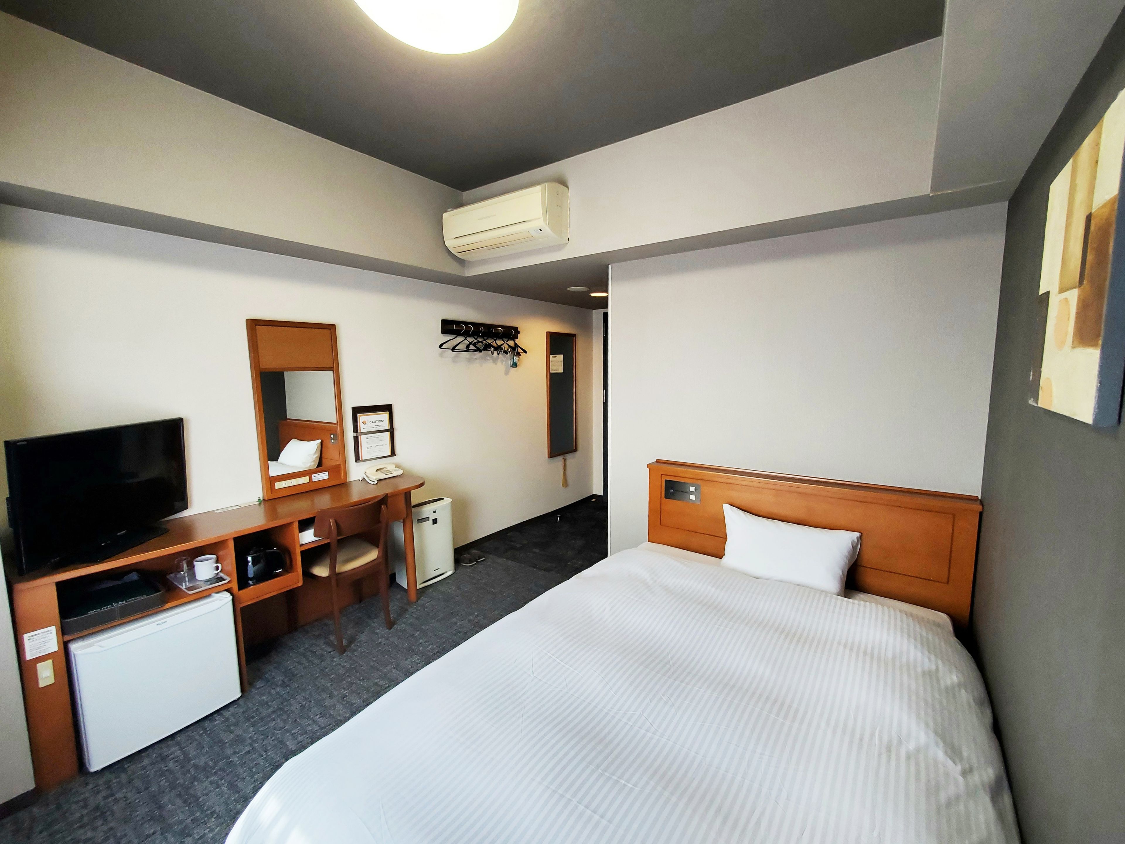 Habitación de hotel con cama, escritorio con televisor, refrigerador pequeño, espejo y equipo de aire acondicionado.