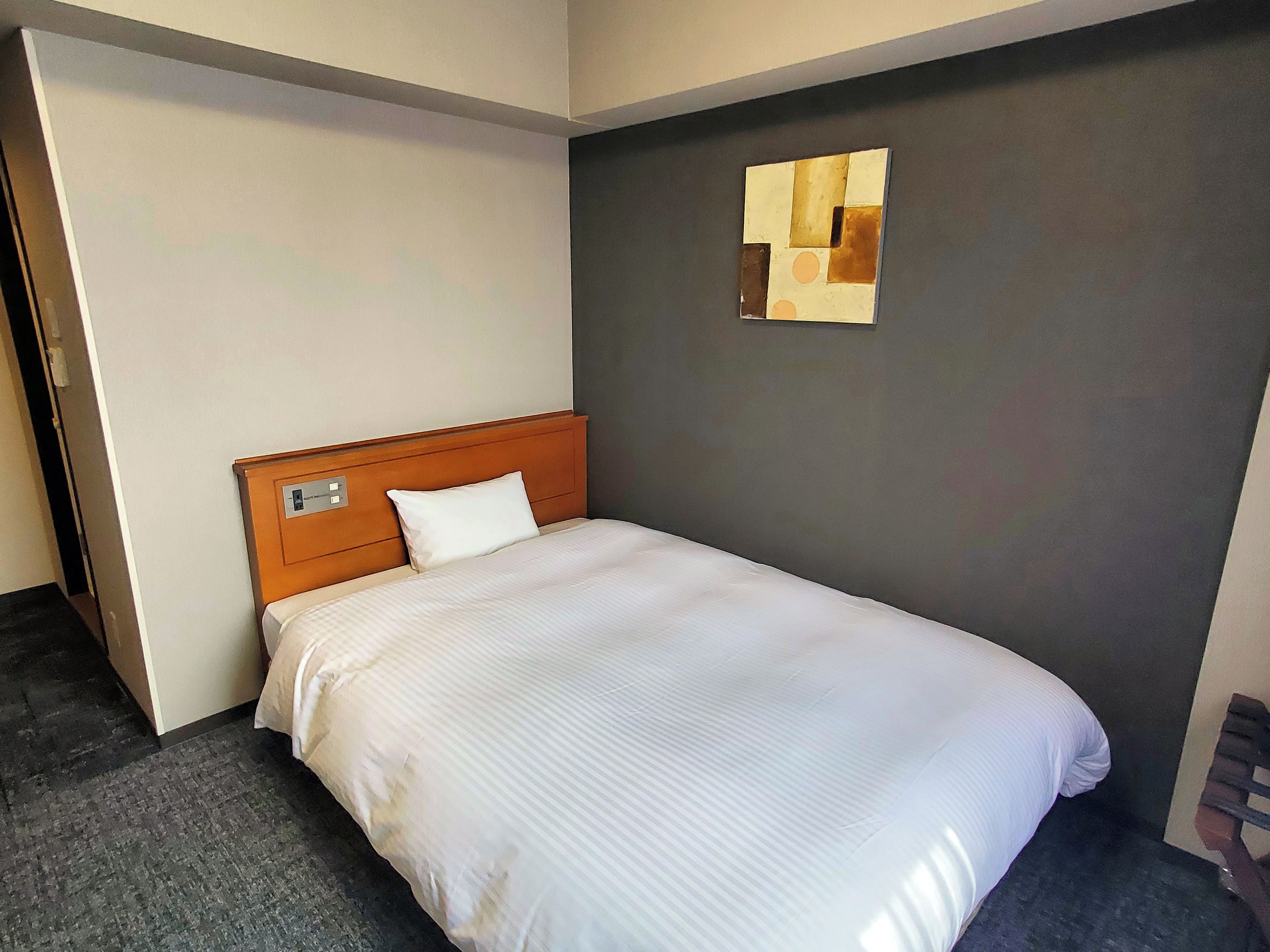 Ein Hotelzimmer mit einem Einzelbett, einem Holzkopfstück, einem grauen Teppich und einem an der Wand befestigten Kunstwerk.
