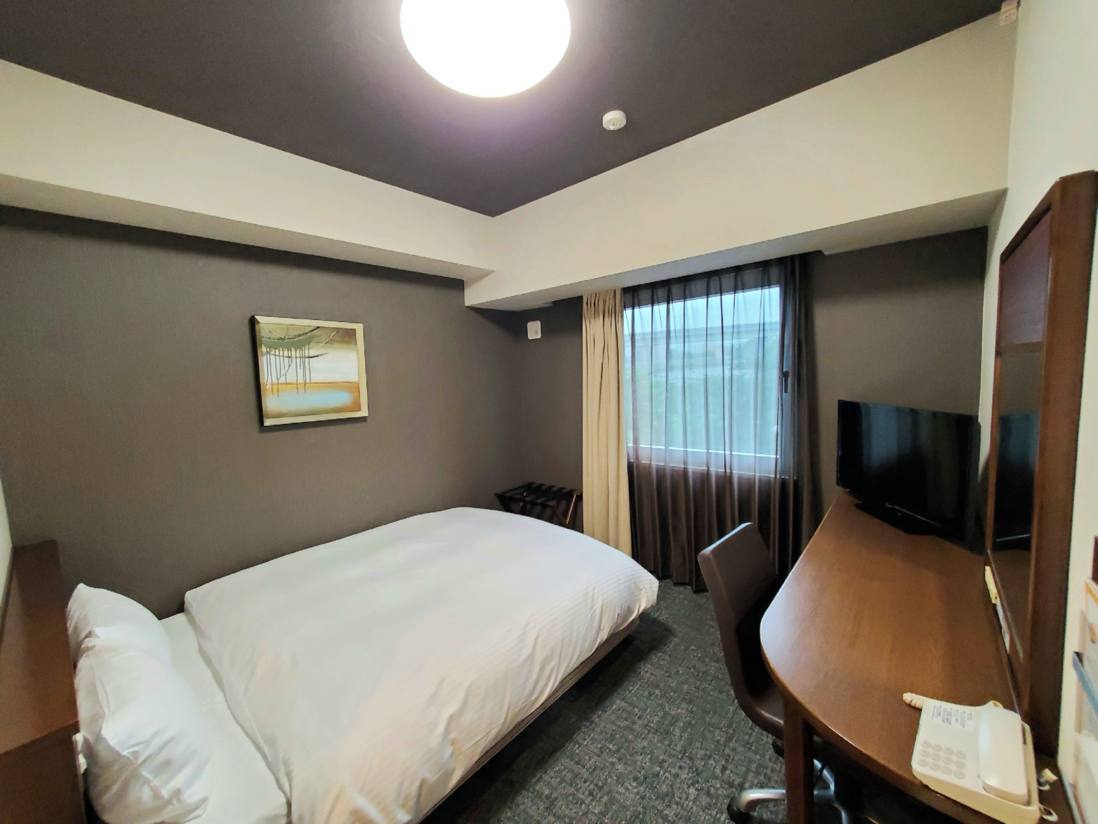 Una camera d'albergo con un letto, una scrivania con un telefono, una televisione, una finestra con tende e un quadro appeso al muro.