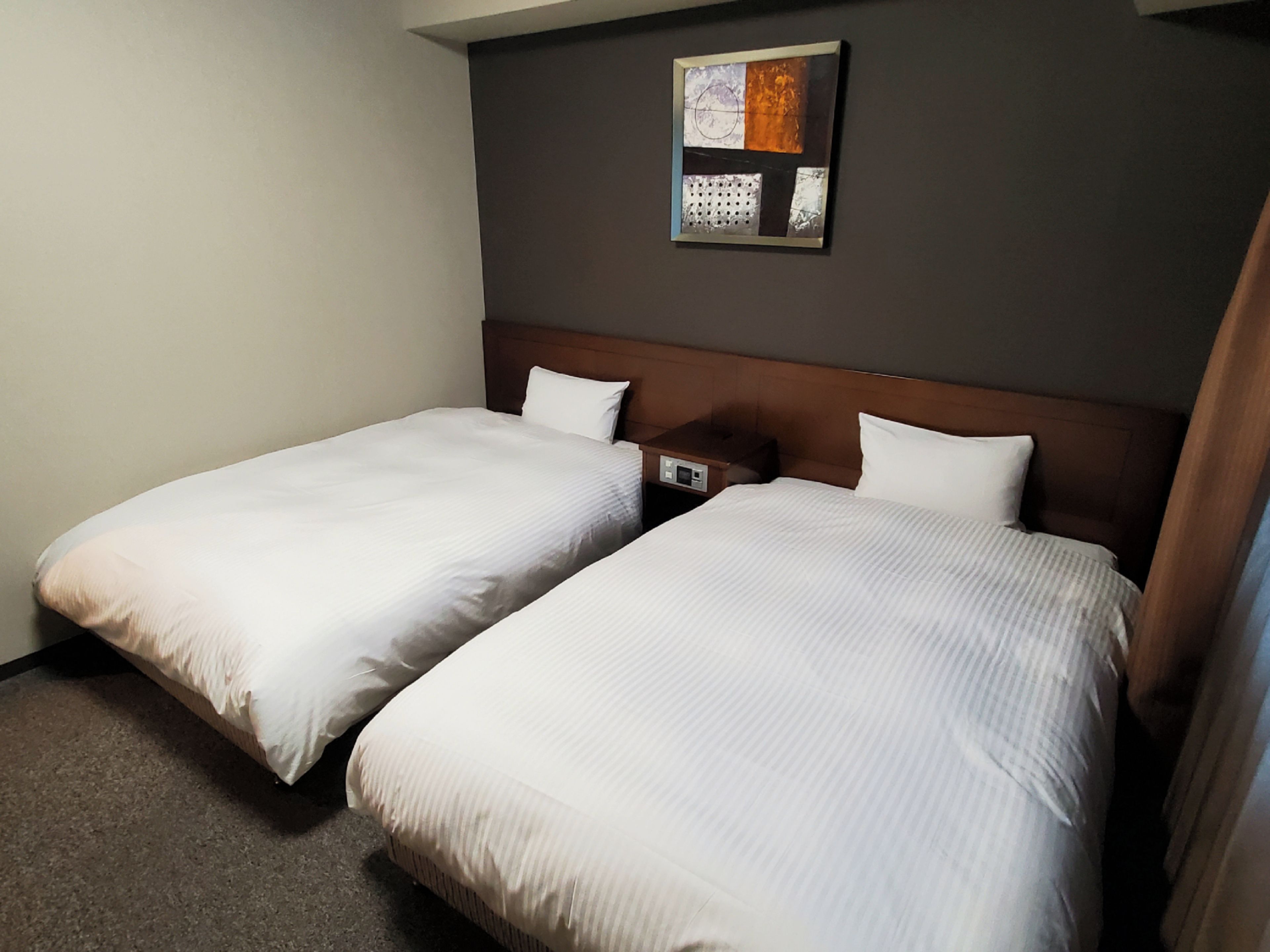 A hotel room with two single beds, a wooden headboard, a small bedside table, and a wall-mounted artwork.