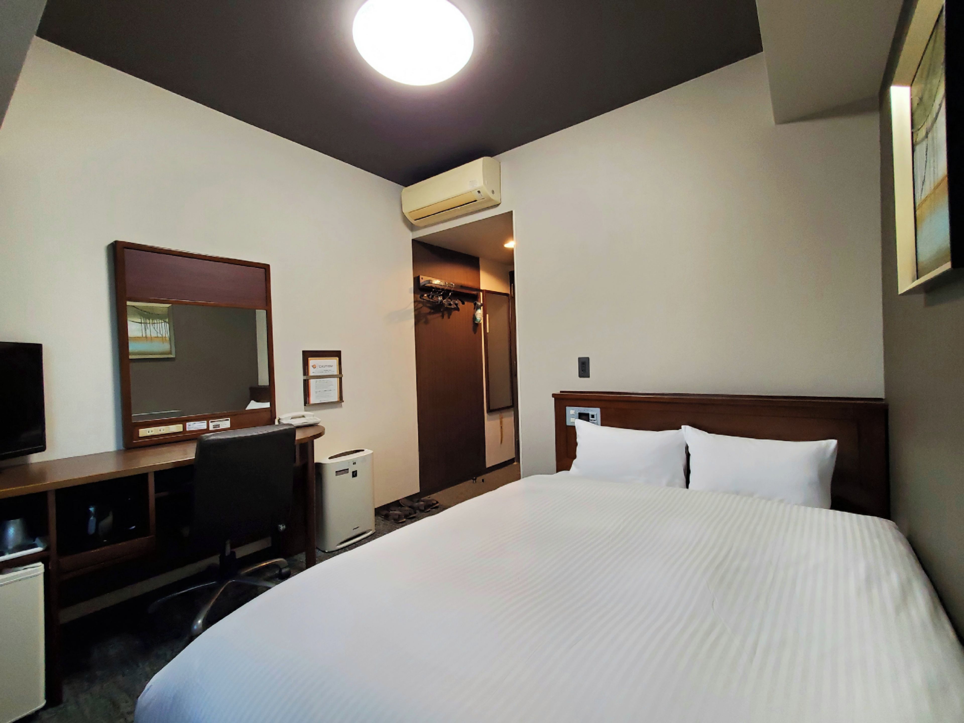 A hotel room with a bed, a desk, a chair, a television, an air conditioning unit, and a mirror.