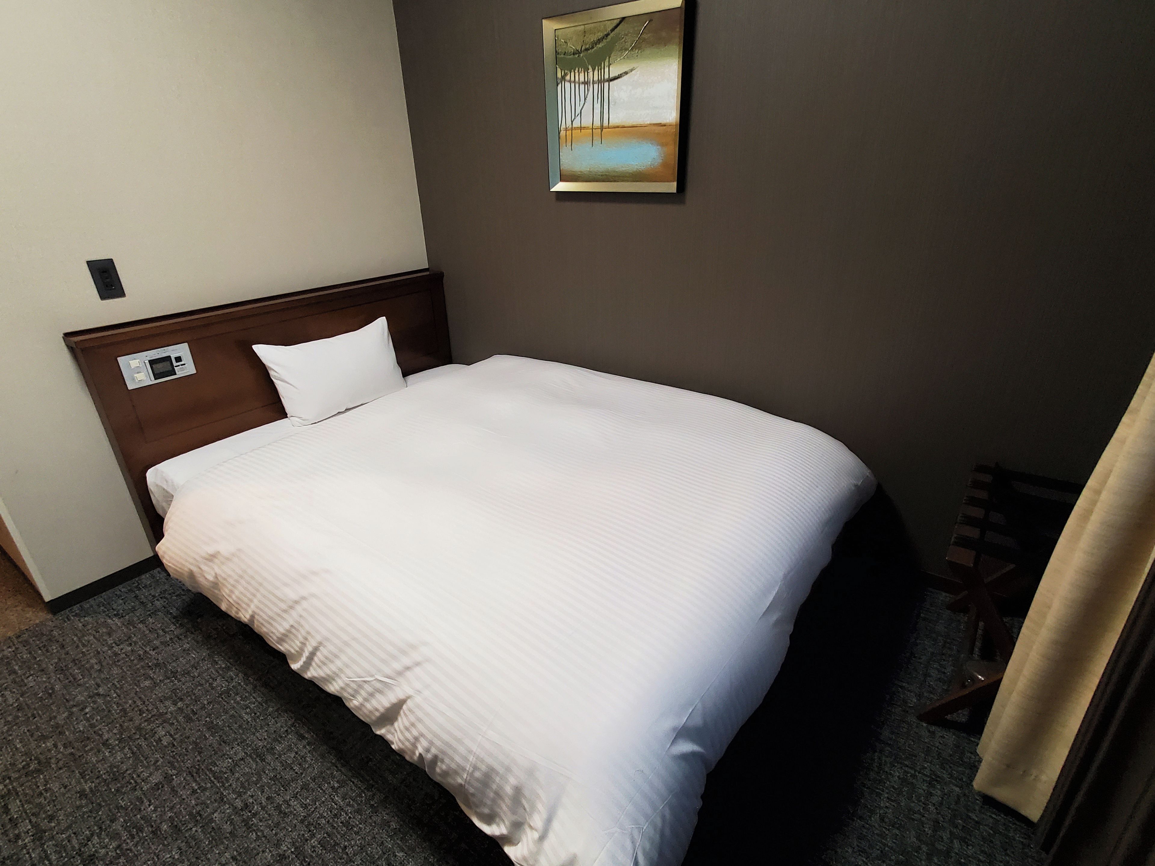 A hotel room with a single bed, a headboard with a control panel, a painting on the wall, and a carpeted floor.