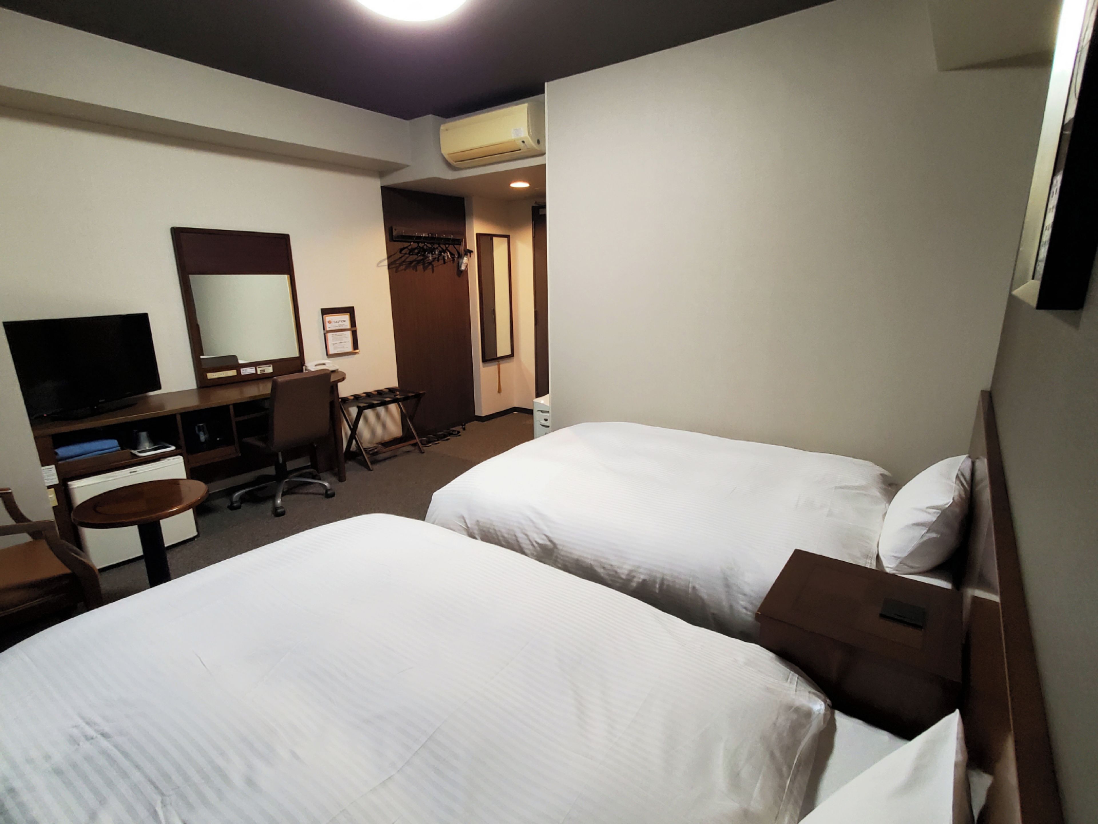 Habitación de hotel con dos camas, un escritorio con una silla, un televisor y un equipo de aire acondicionado.
