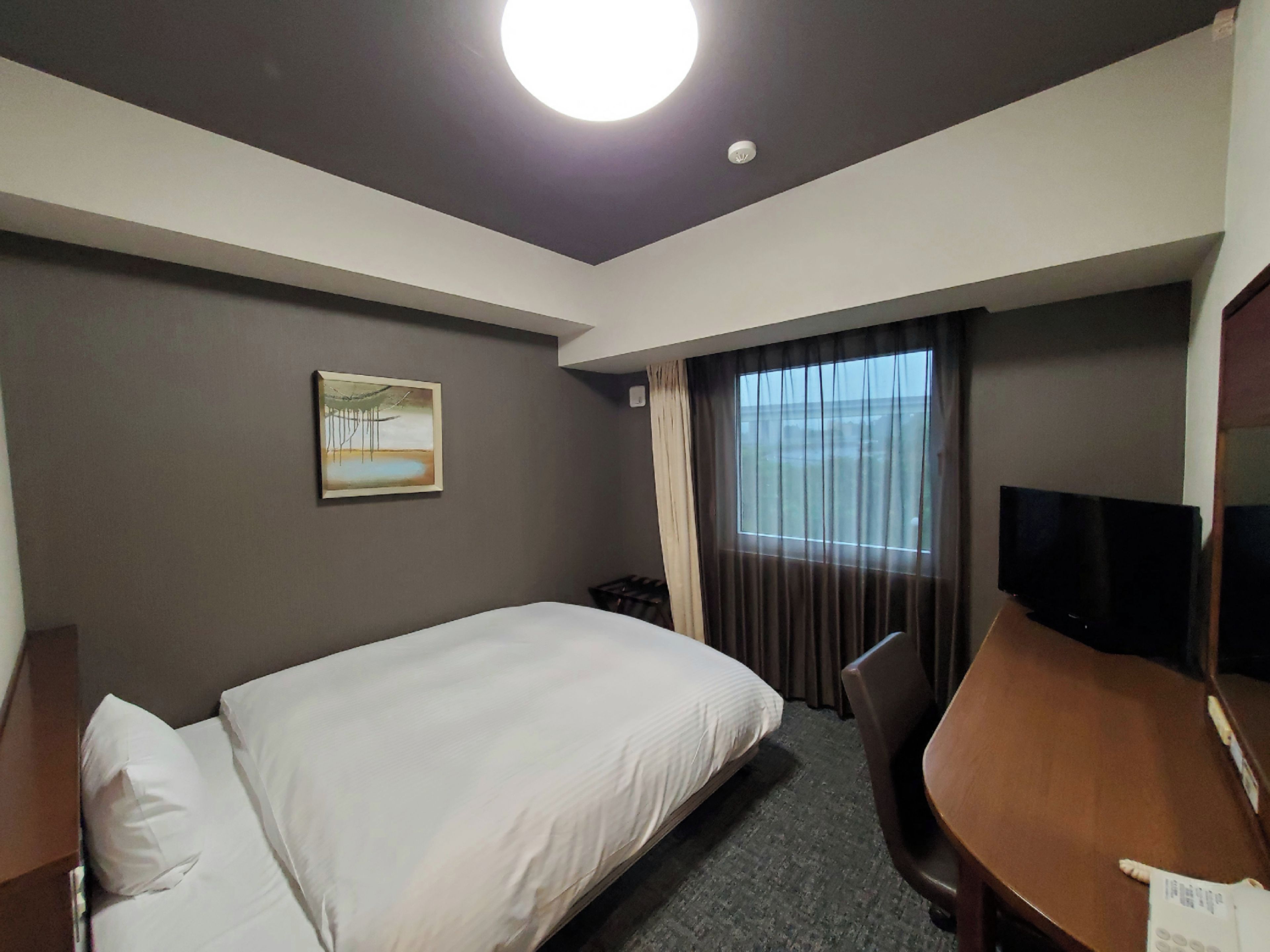 A hotel room with a bed, a desk, a chair, a television, a painting on the wall, and a window with curtains.