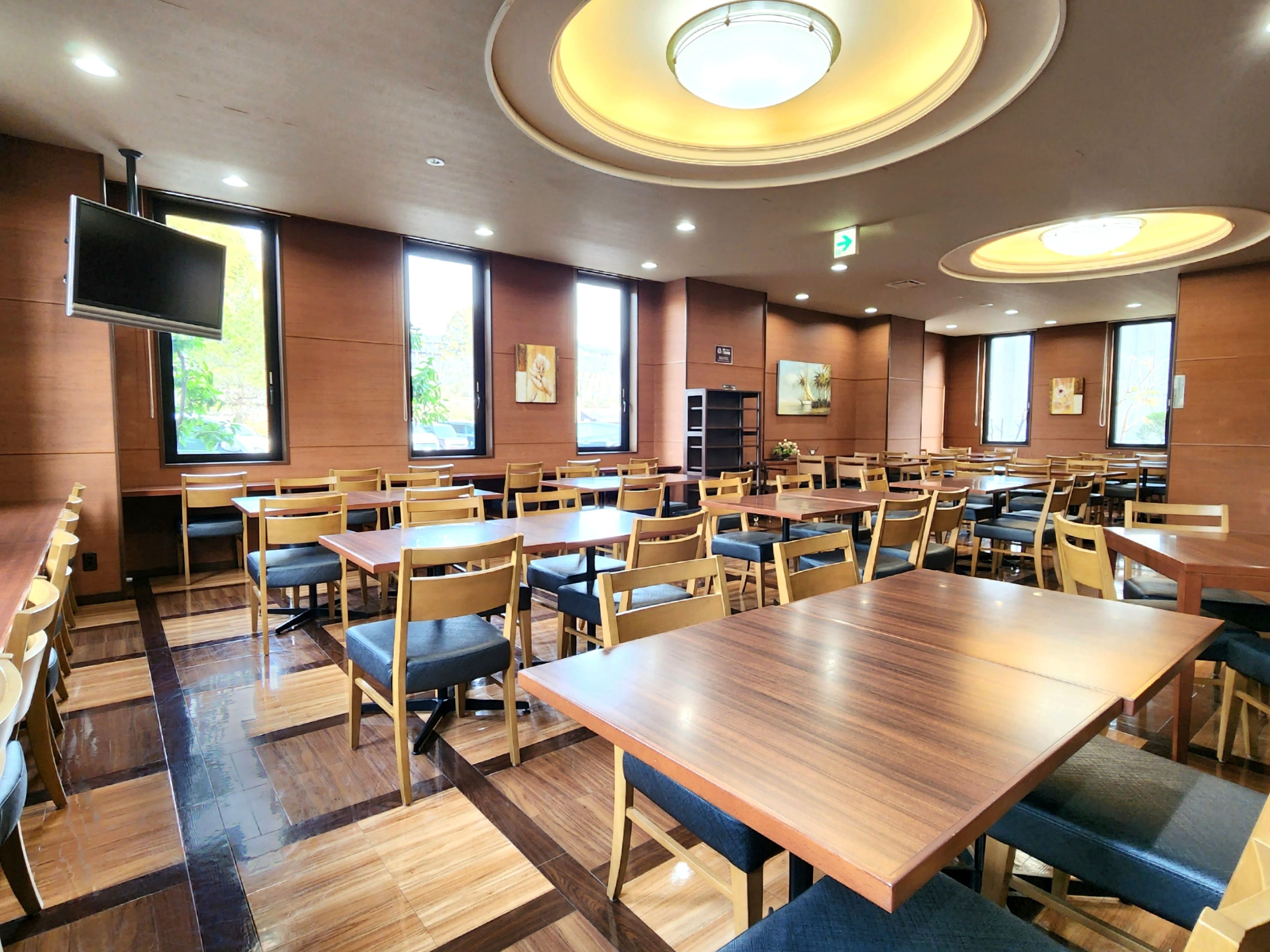 A spacious dining area with multiple tables and chairs, large windows, and a flat-screen TV mounted on the wall.