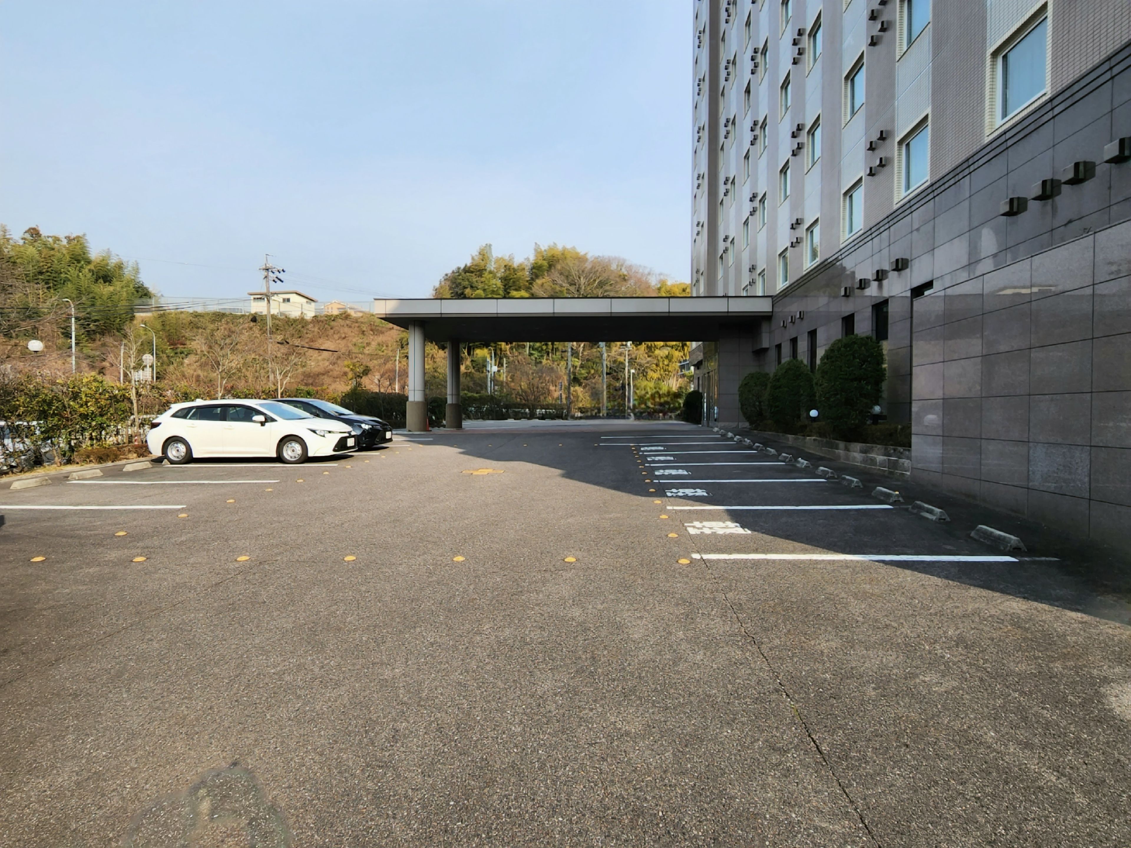A parking lot with a white car parked, a building with a flat roof, and trees in the background.