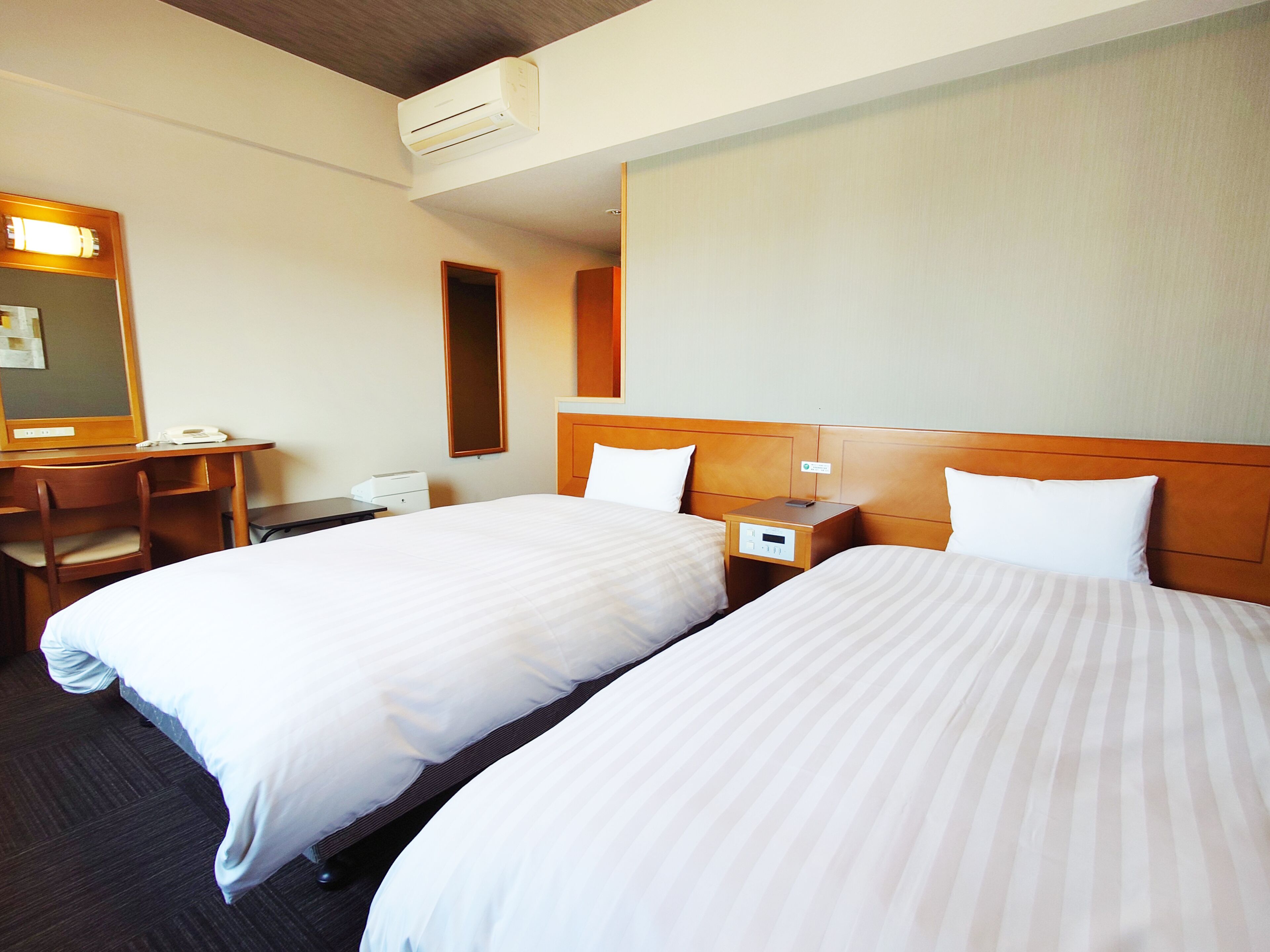 A hotel room with two beds, a desk, a chair, a nightstand, and an air conditioning unit.