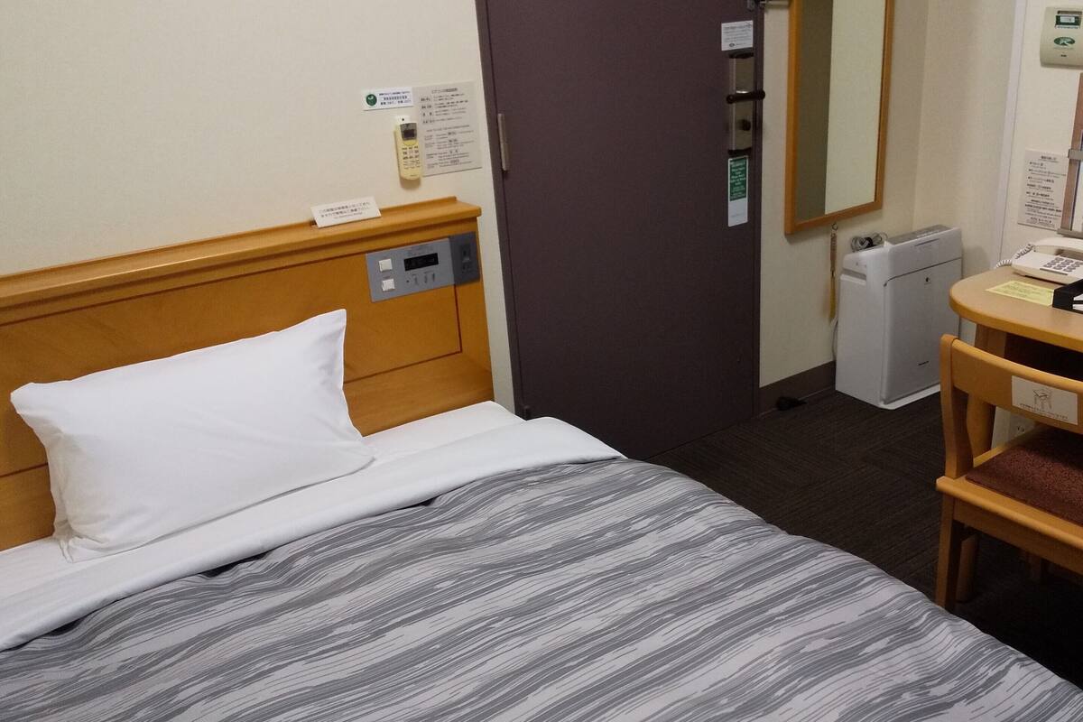 Ein Hotelzimmer mit einem Bett, einem Schreibtisch mit Stuhl, einem Spiegel, einem Thermostat und einem kleinen Kühlschrank.