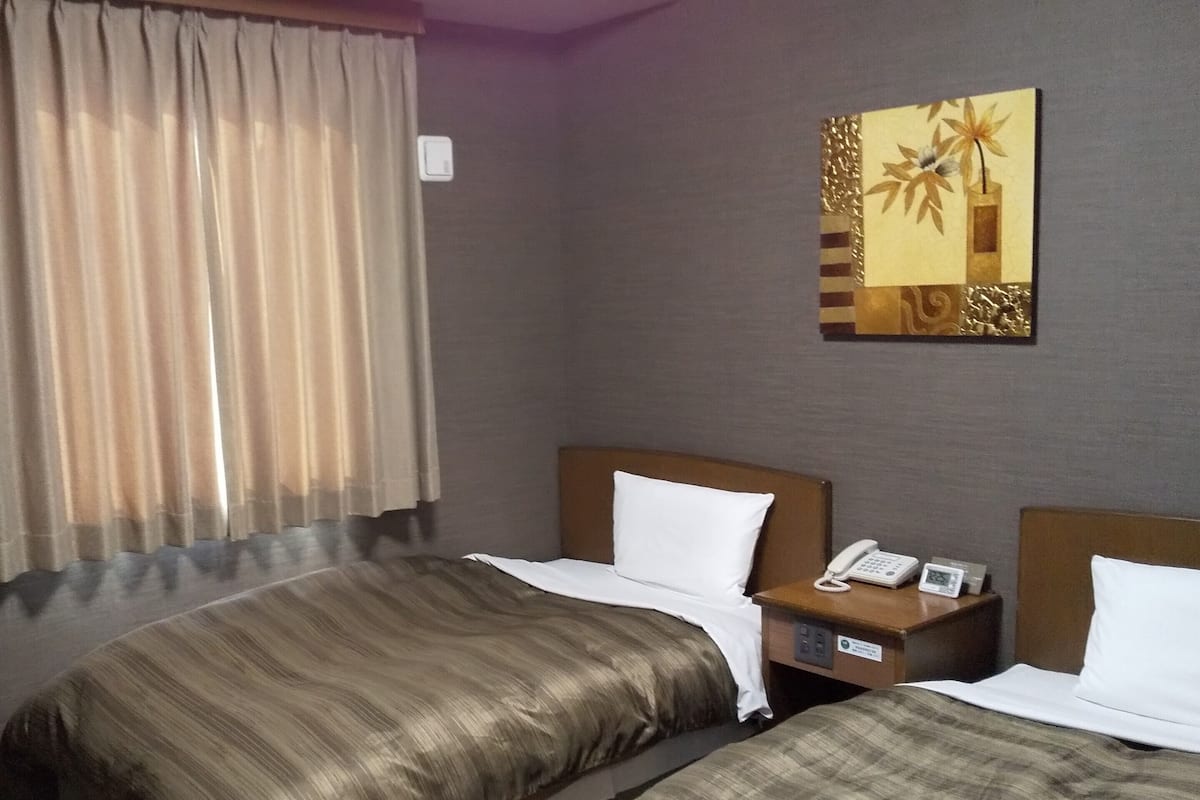 Ein Hotelzimmer mit zwei Betten, einem Nachttisch mit Telefon und Uhr, einem Bild an der Wand und einem Fenster mit Vorhängen.
