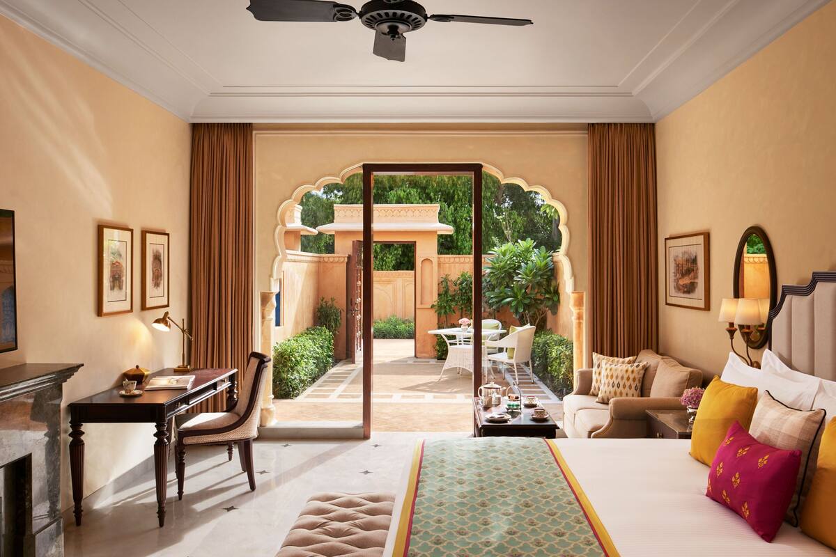 A hotel room with a bed, a desk, a chair, a table with a lamp, and a view of an outdoor garden through an open door.