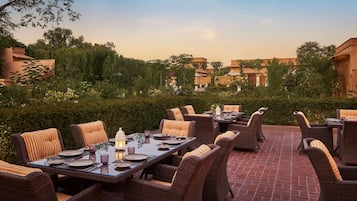 An outdoor dining area with wicker furniture and a brick floor, surrounded by greenery and buildings in the background.