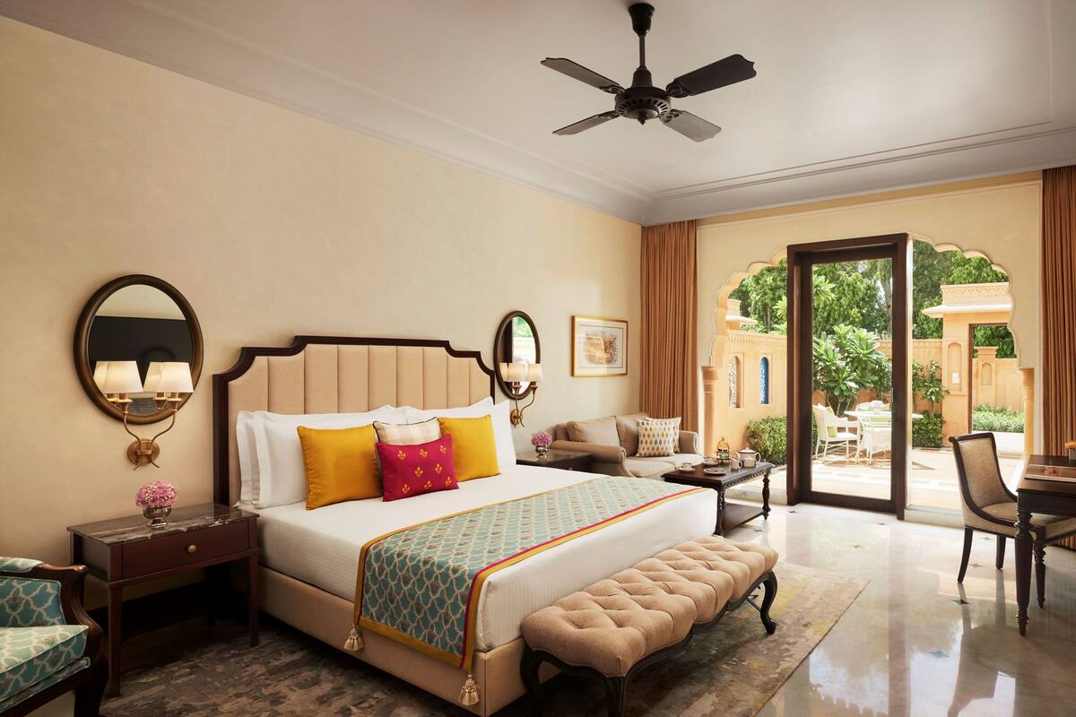 A spacious bedroom with a large bed, a sitting area with a sofa and armchairs, a dining table, and a view of the outdoor garden through a glass door.