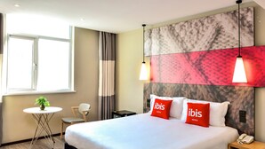 Une chambre d’hôtel avec un grand lit, deux oreillers rouges portant la marque « ibis hotels », une petite table ronde avec une plante, une chaise et une table de chevet avec un téléphone.