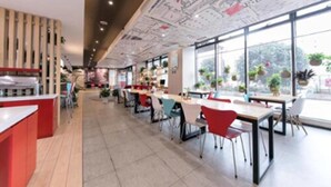 Un restaurant moderne avec un long comptoir, de nombreuses tables avec des chaises et de grandes fenêtres ornées de plantes.