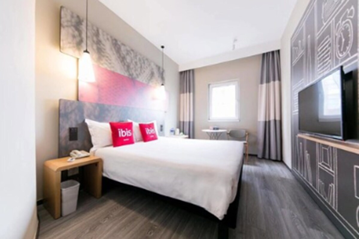 Une chambre d’hôtel moderne avec un grand lit, une table de chevet avec une lampe, une télévision et une fenêtre avec des rideaux.