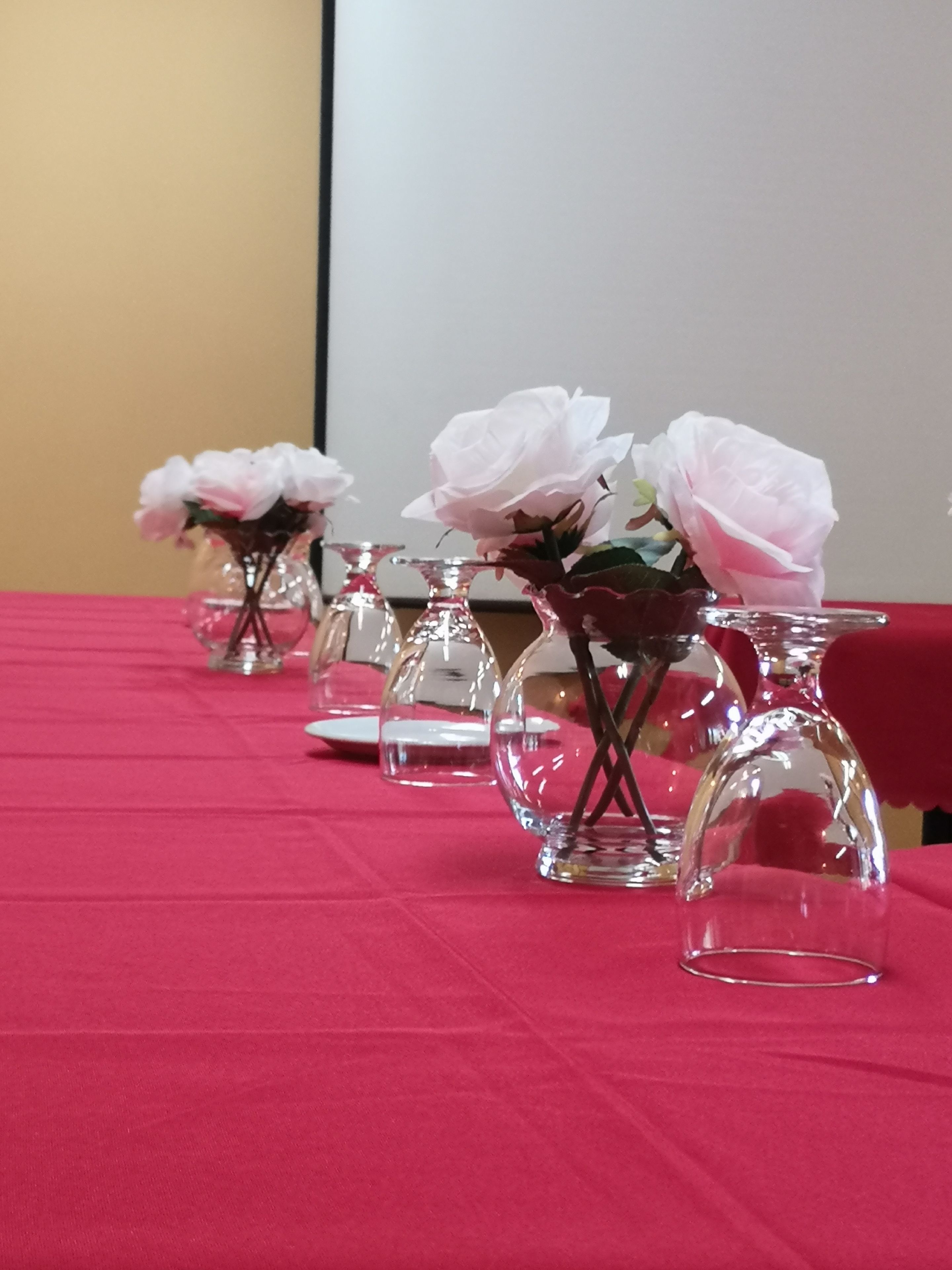 Een tafel met glazen vaasjes met witte bloemen, op een rode tafelkleed gezet.