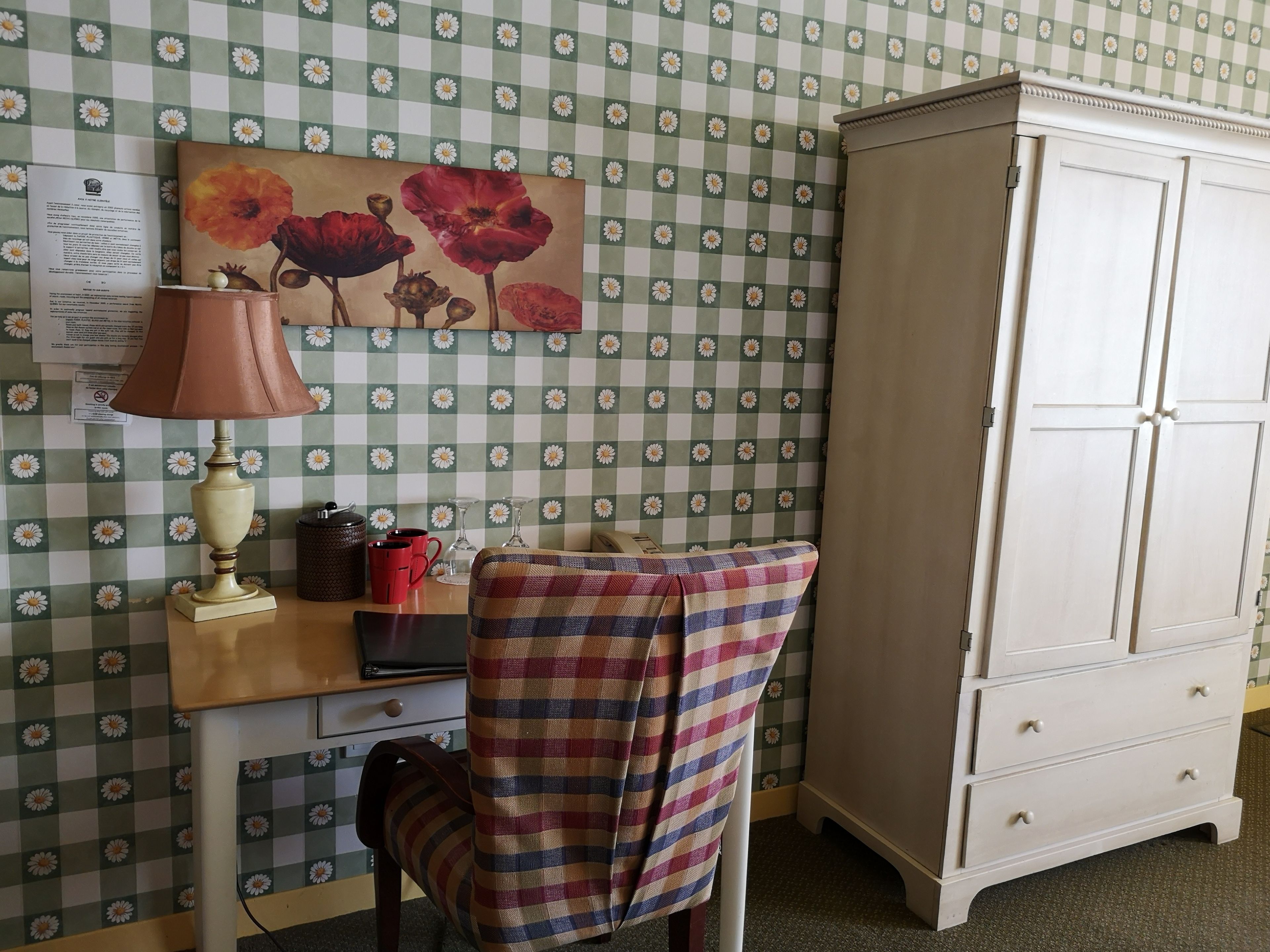 Une pièce avec un papier peint à motifs floraux à carreaux, un bureau avec une lampe, une chaise recouverte d’un tissu à carreaux et une armoire blanche.