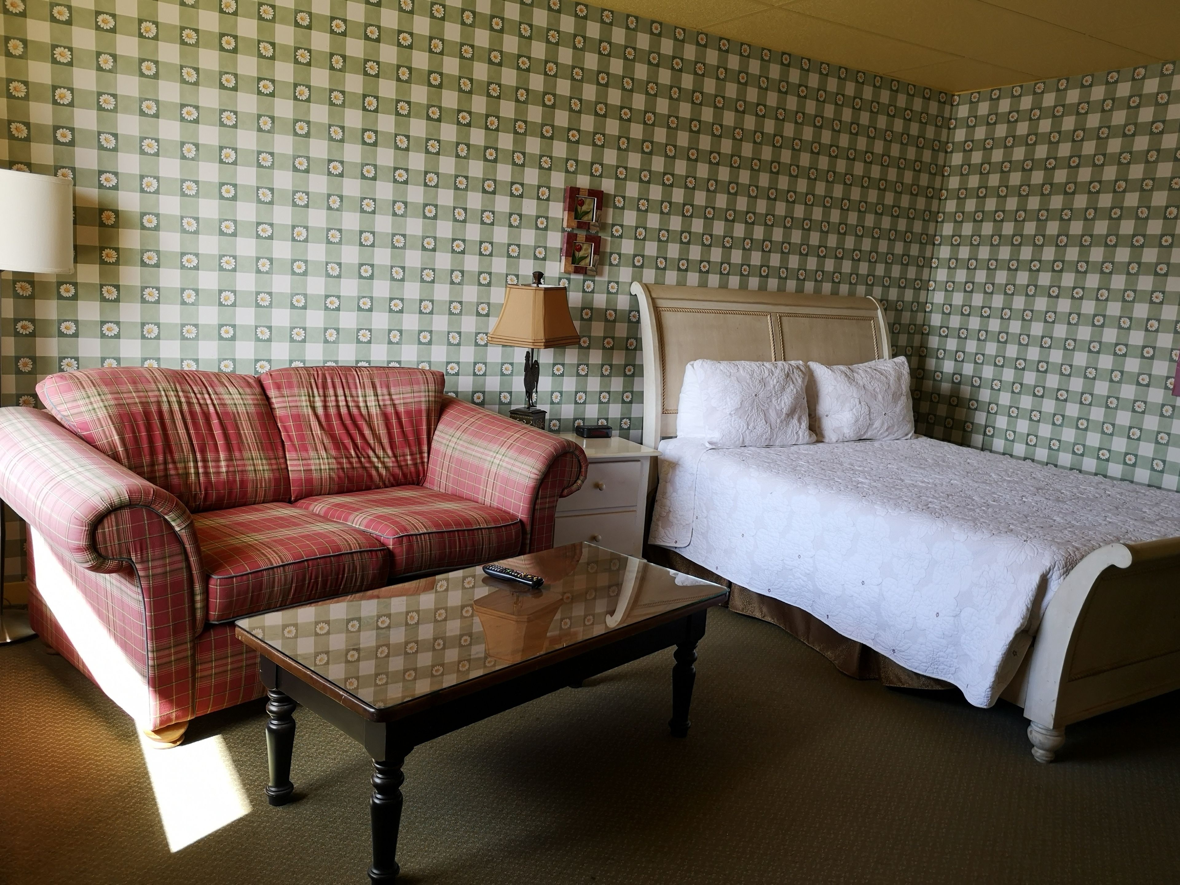A room with a checkered floral wallpaper, a plaid sofa, a single bed with a white quilt, a bedside table, and a lamp.