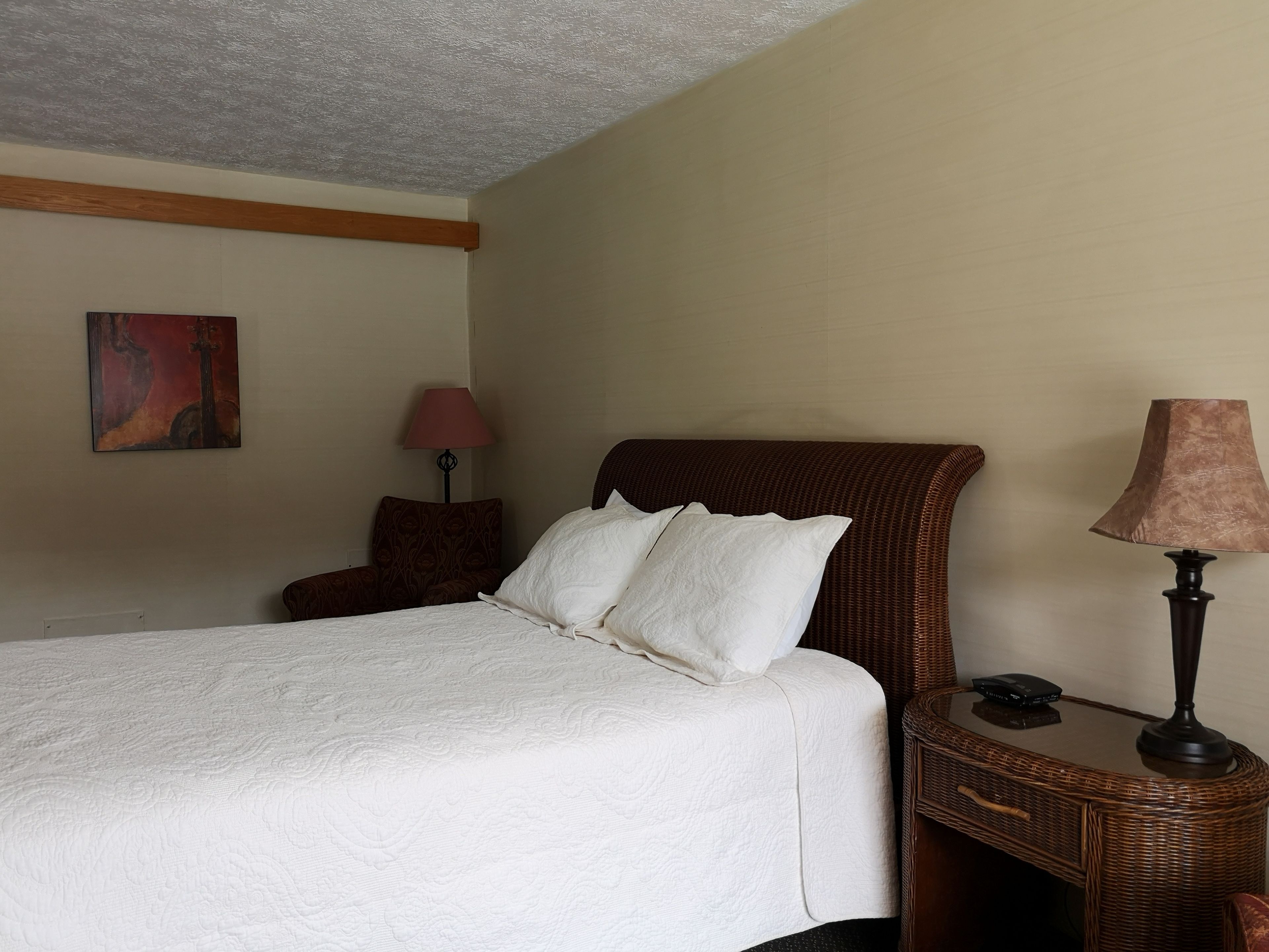 A neatly made bed with white linens, a wicker headboard, and a bedside table with a lamp and a phone.