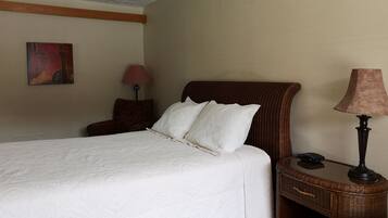A neatly made bed with white linens, a wicker headboard, and a bedside table with a lamp and a phone.