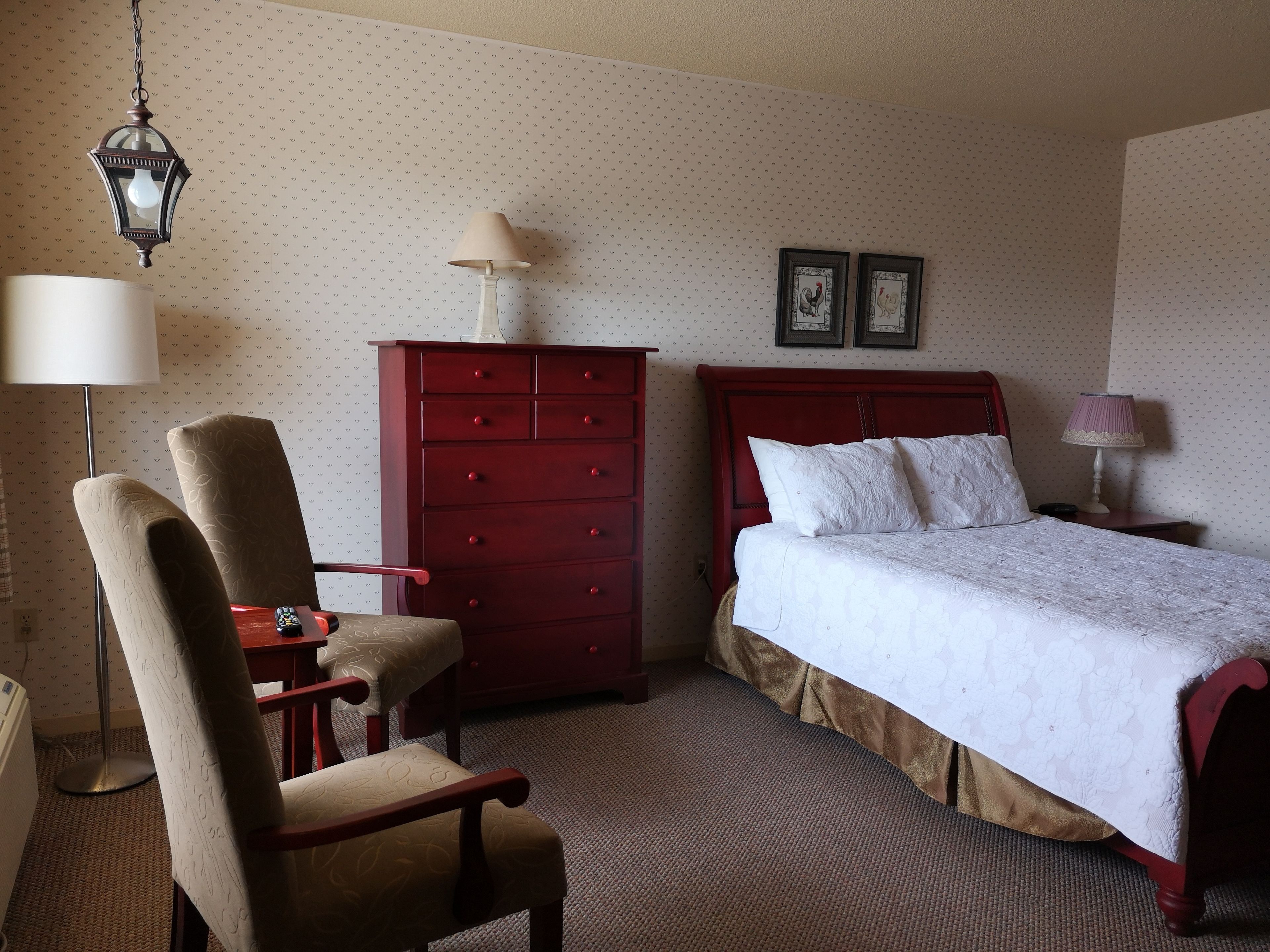 A hotel room with a bed, a dresser, a chair, a lamp, and two framed pictures on the wall.
