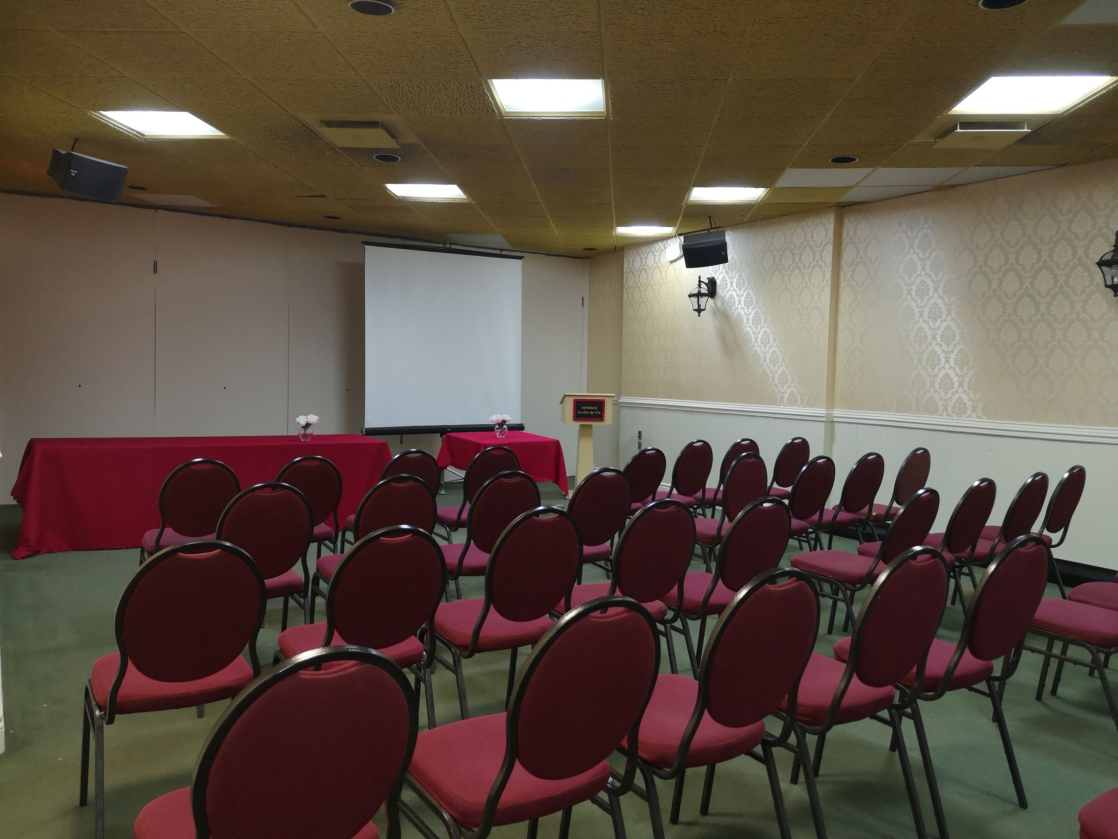 A conference room with rows of red chairs, a projector screen, and a red table.