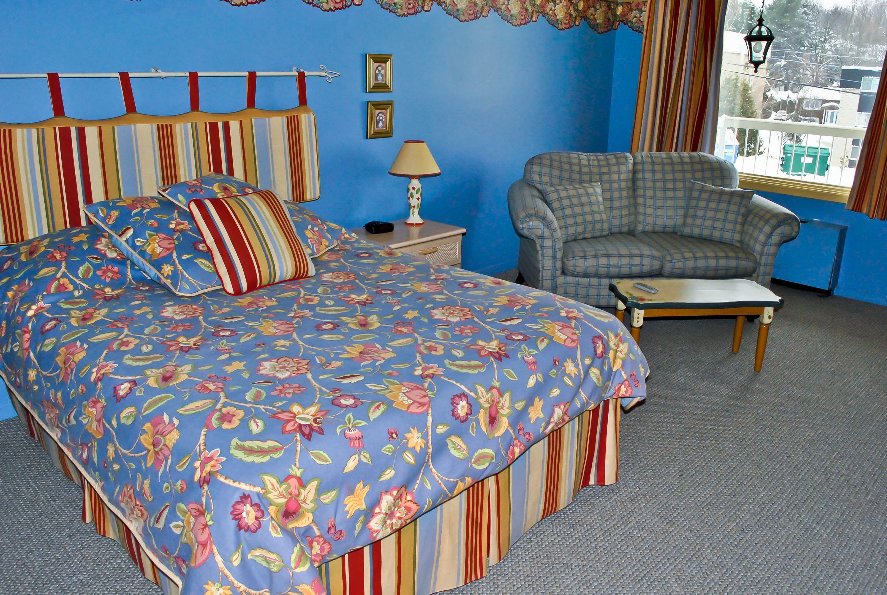 Um quarto com cabeceira listrada, colcha florida, poltrona xadrez e uma mesa pequena.