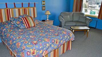 Um quarto com cabeceira listrada, colcha florida, poltrona xadrez e uma mesa pequena.