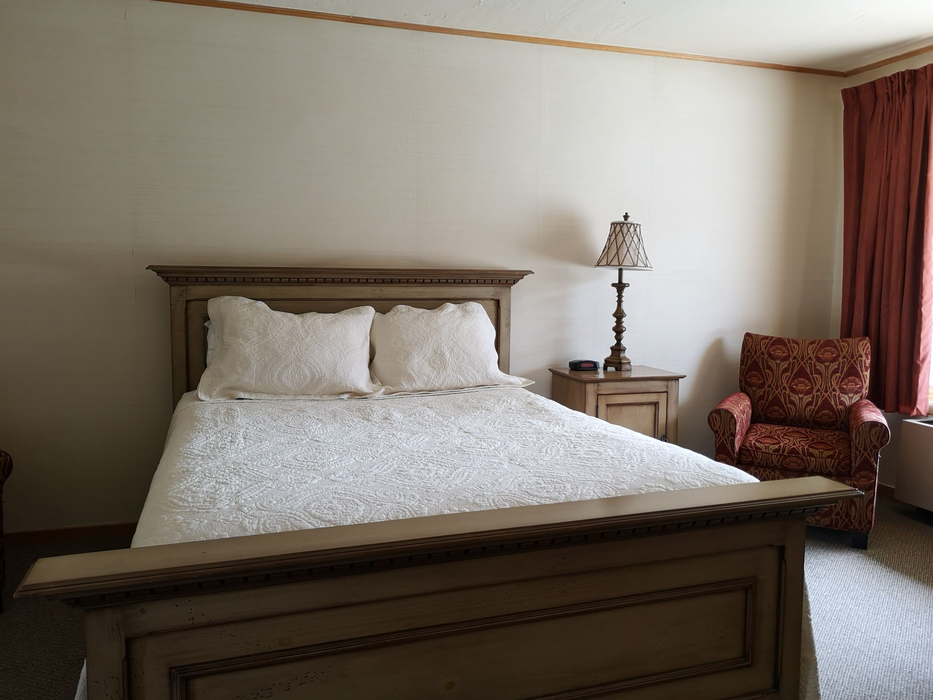 Uma cama bem arrumada com cabeceira de madeira, uma mesa de cabeceira com abajur e uma poltrona estampada.