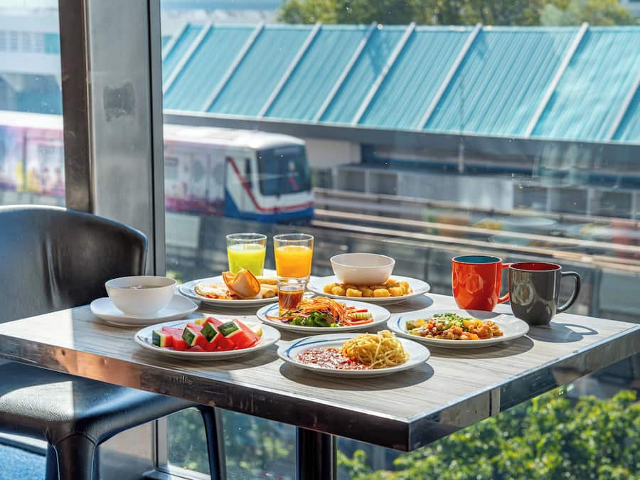 Una mesa de desayuno con platos de comida, un bol y dos tazas, con vistas a un tren y tejados verdes.