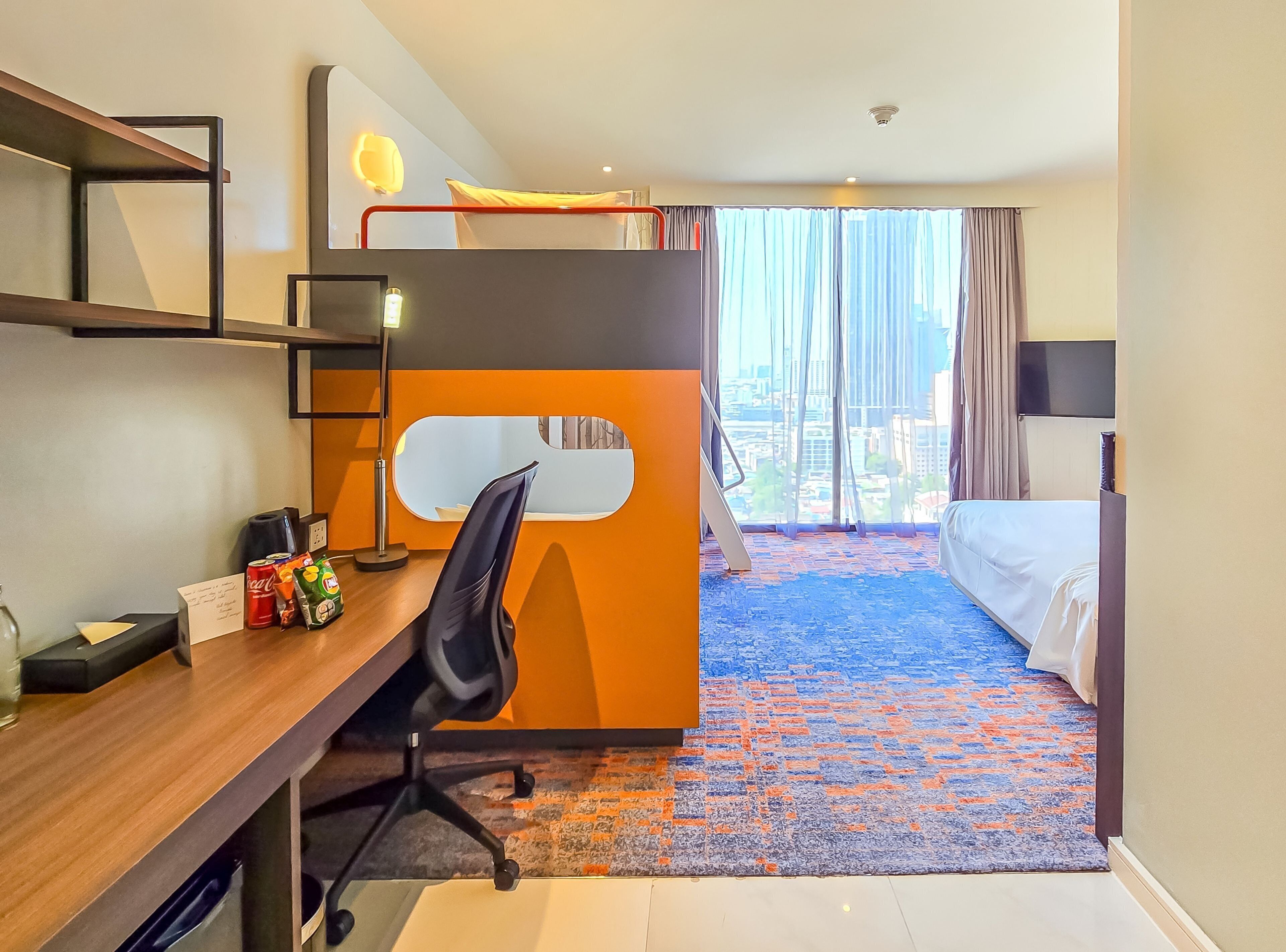 A hotel room with a bunk bed, desk, chair, and a large window with a city view.