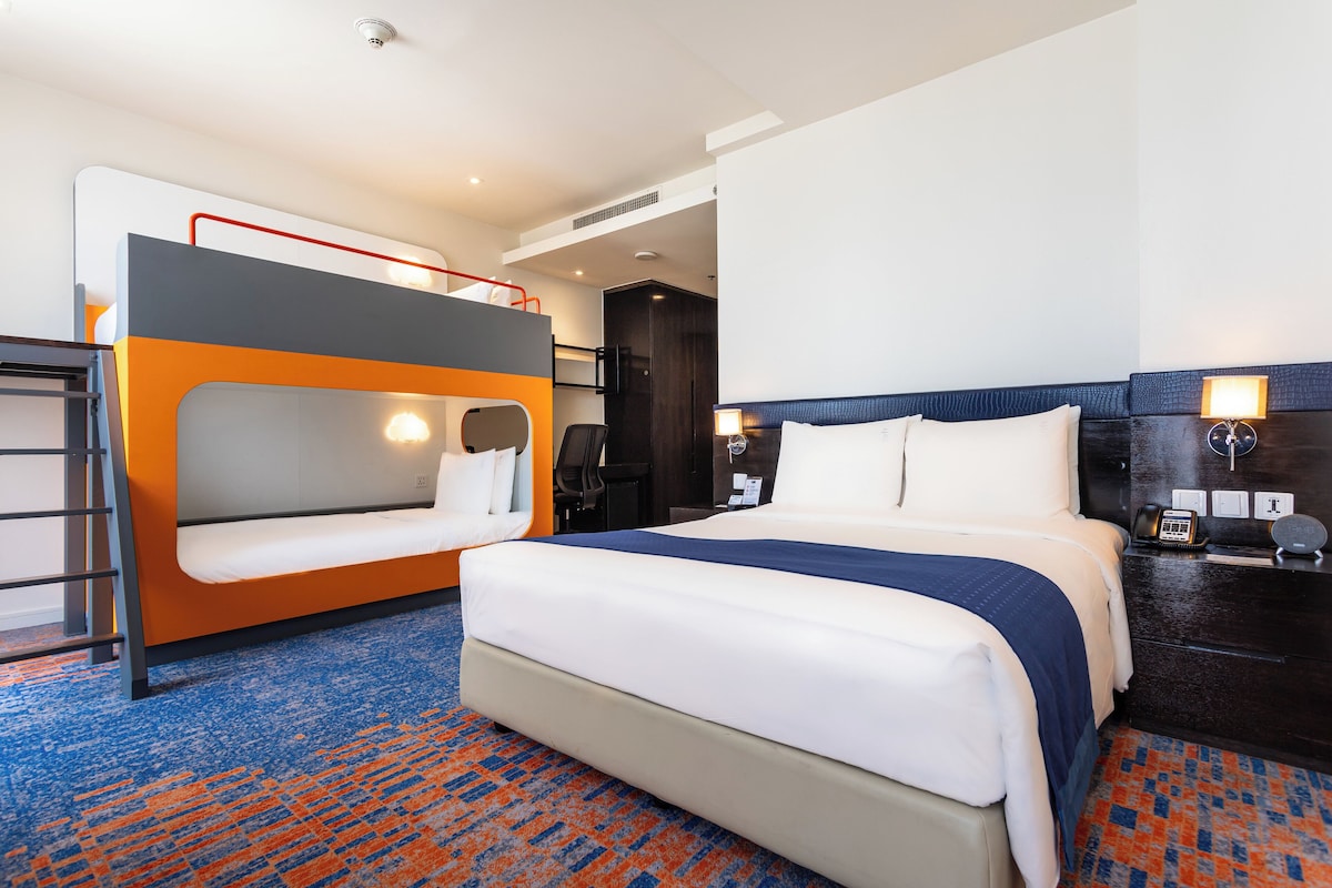 Habitació d'hotel amb una llitera, un llit individual, escriptori amb làmpada i un bany visible al fons.