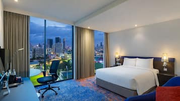 A hotel room with a large bed, a desk with a chair, a TV, and a view of a cityscape.