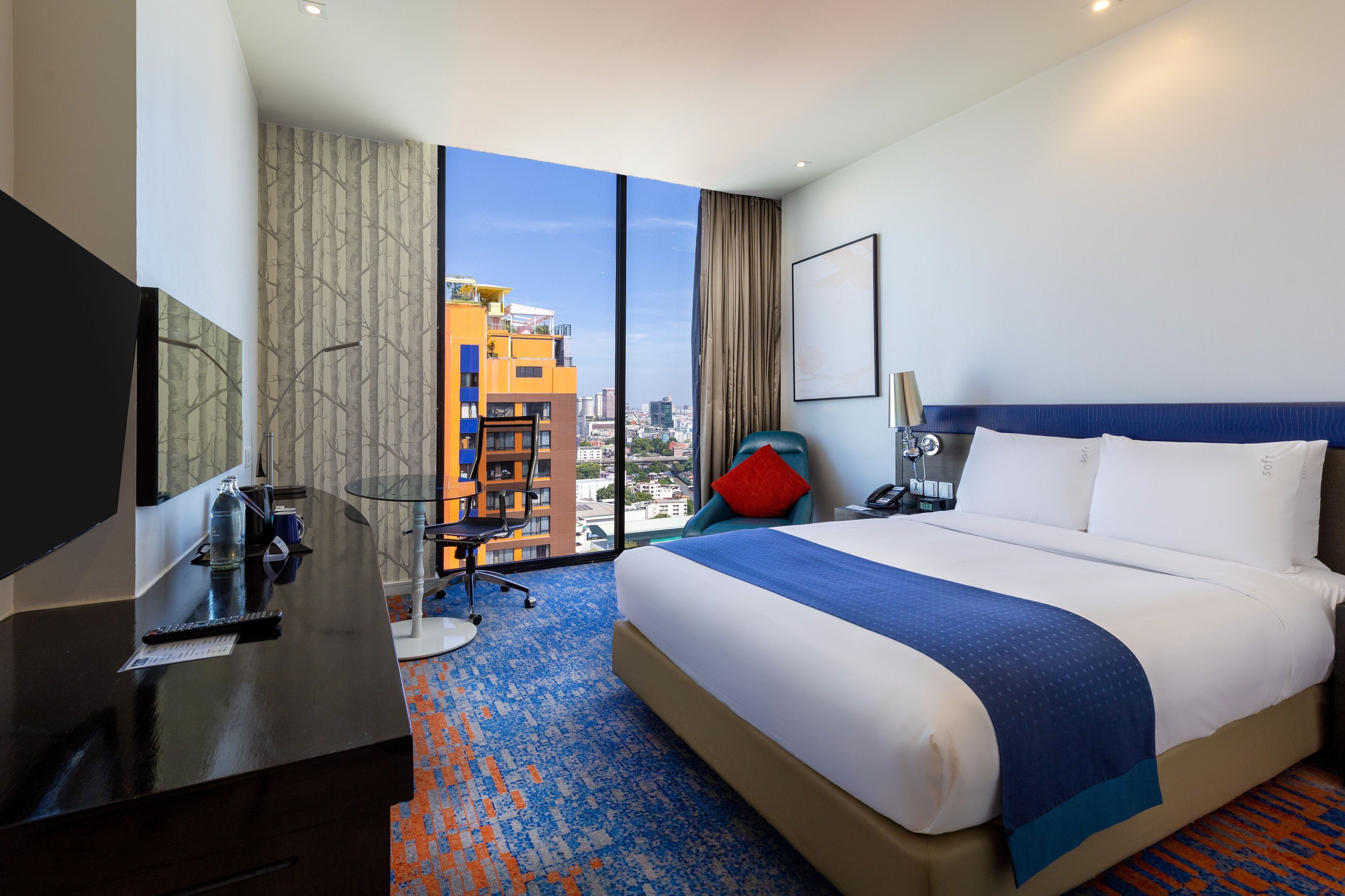 Una habitación de hotel con una cama grande, un escritorio con silla, televisor y vistas a la ciudad a través de amplios ventanales.