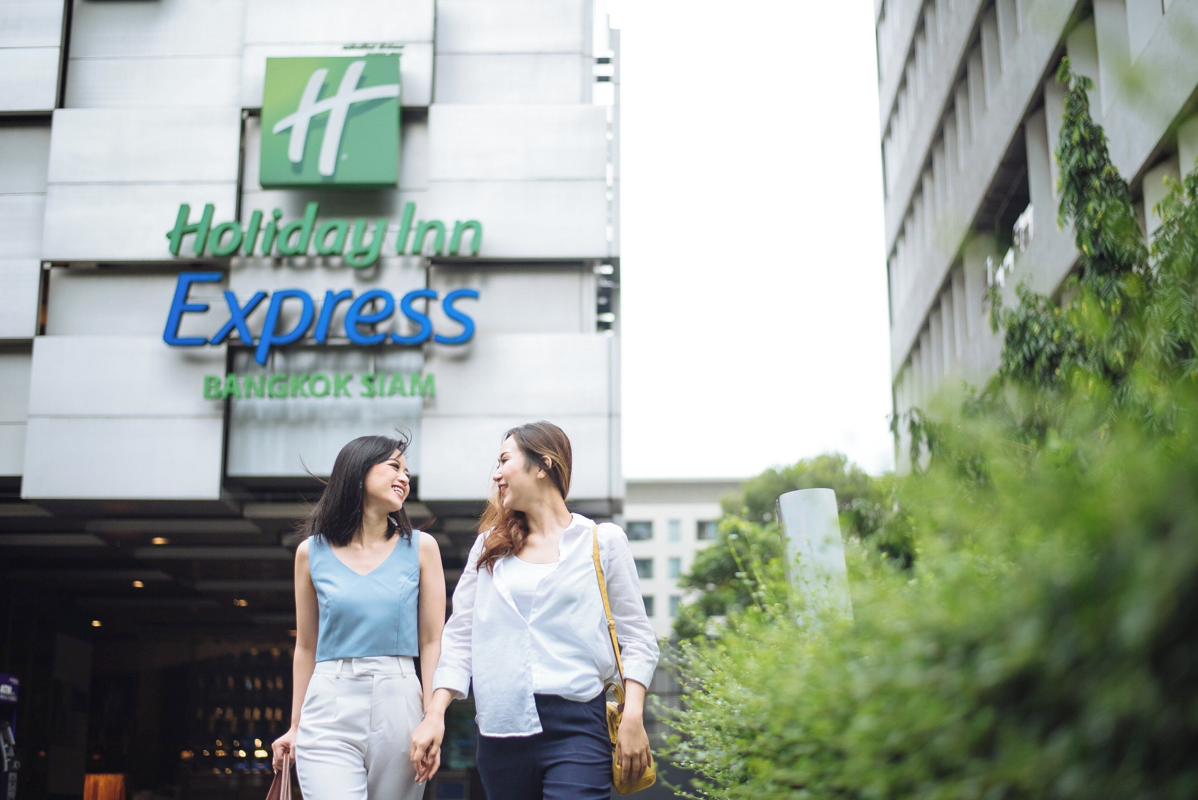 Zwei Personen gehen vor einem Gebäude, an dem ein Schild für Holiday Inn Express Bangkok Siam zu sehen ist.
