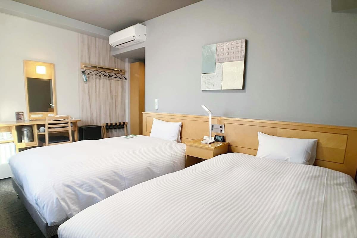 Ett hotellrum med två sängar, ett skrivbord med en stol, en spegel, en luftkonditionering och ett konstverk som hänger på väggen.