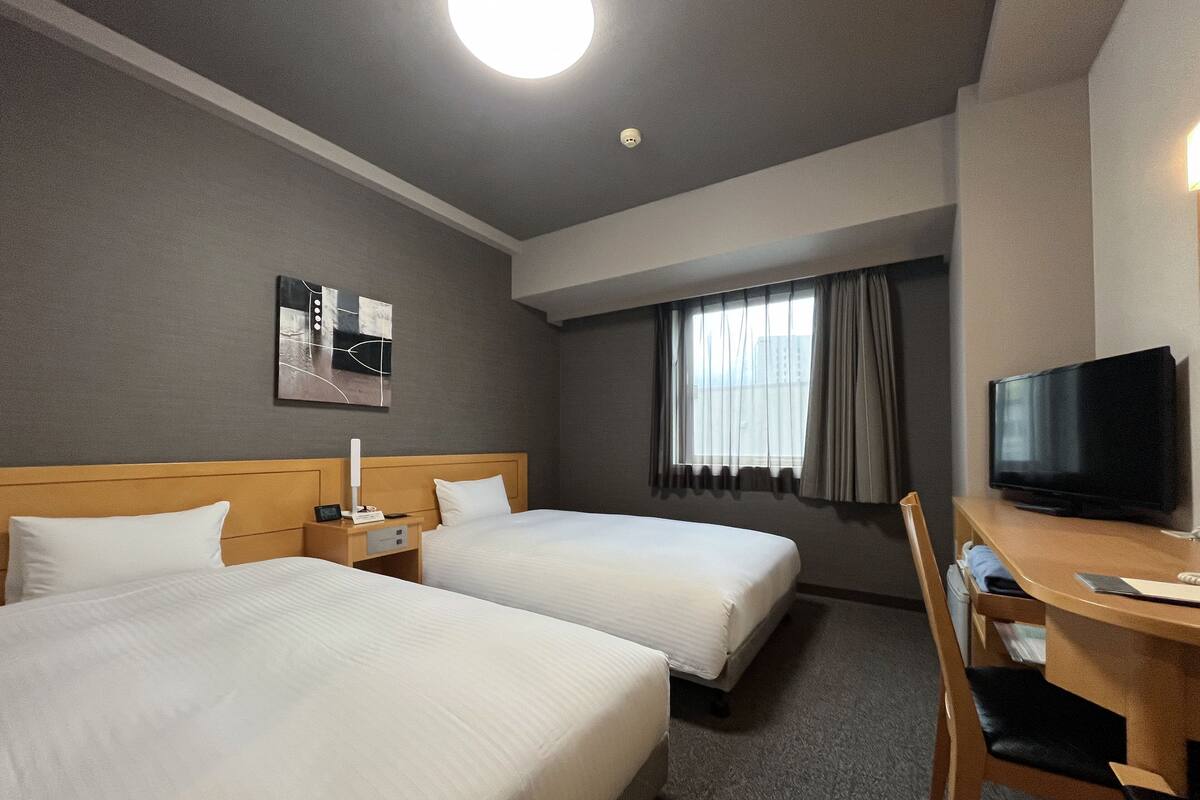 Habitación de hotel con dos camas, un escritorio con silla, televisor y ventana con cortinas.