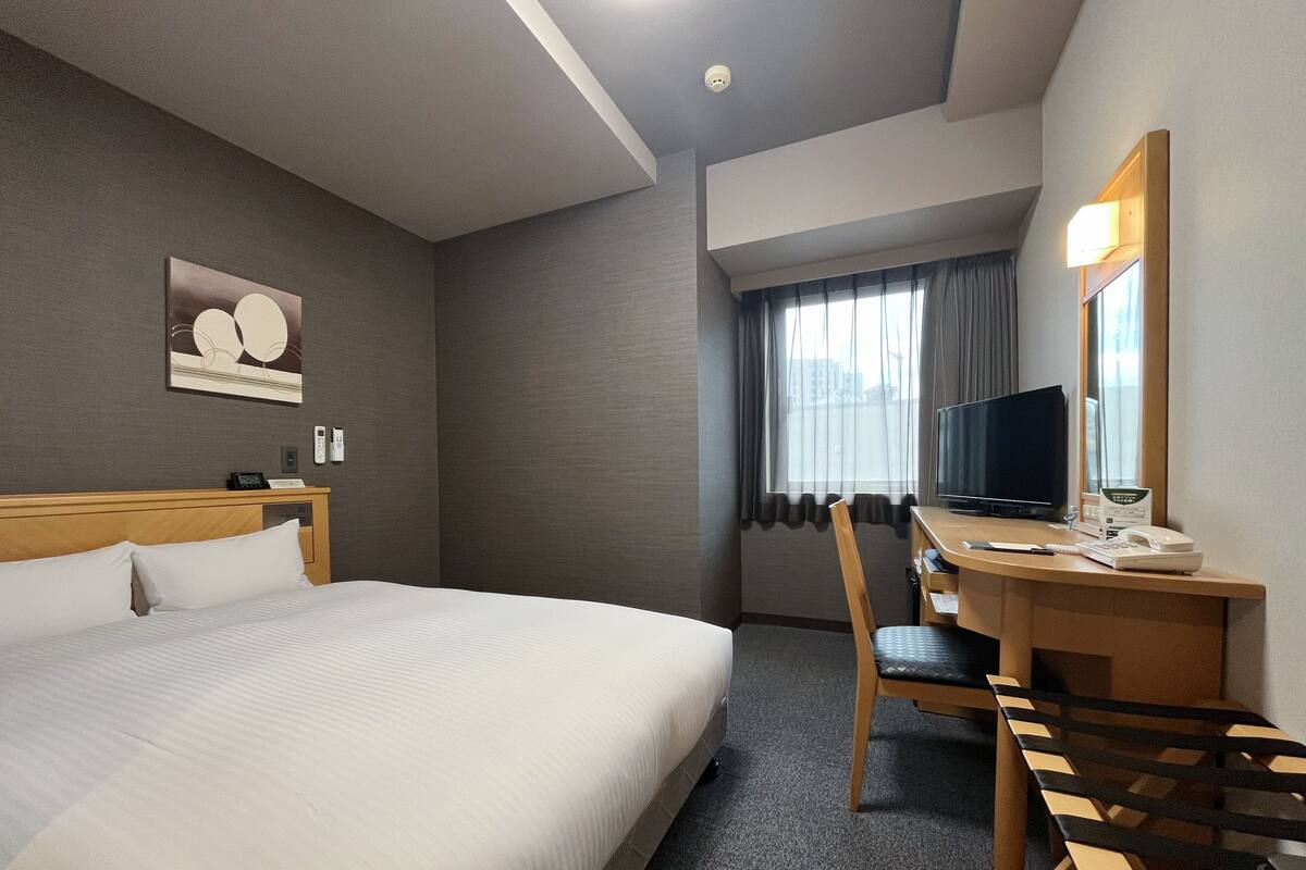 Una habitación de hotel con una cama grande, un escritorio con silla, televisor y ventana con cortinas.
