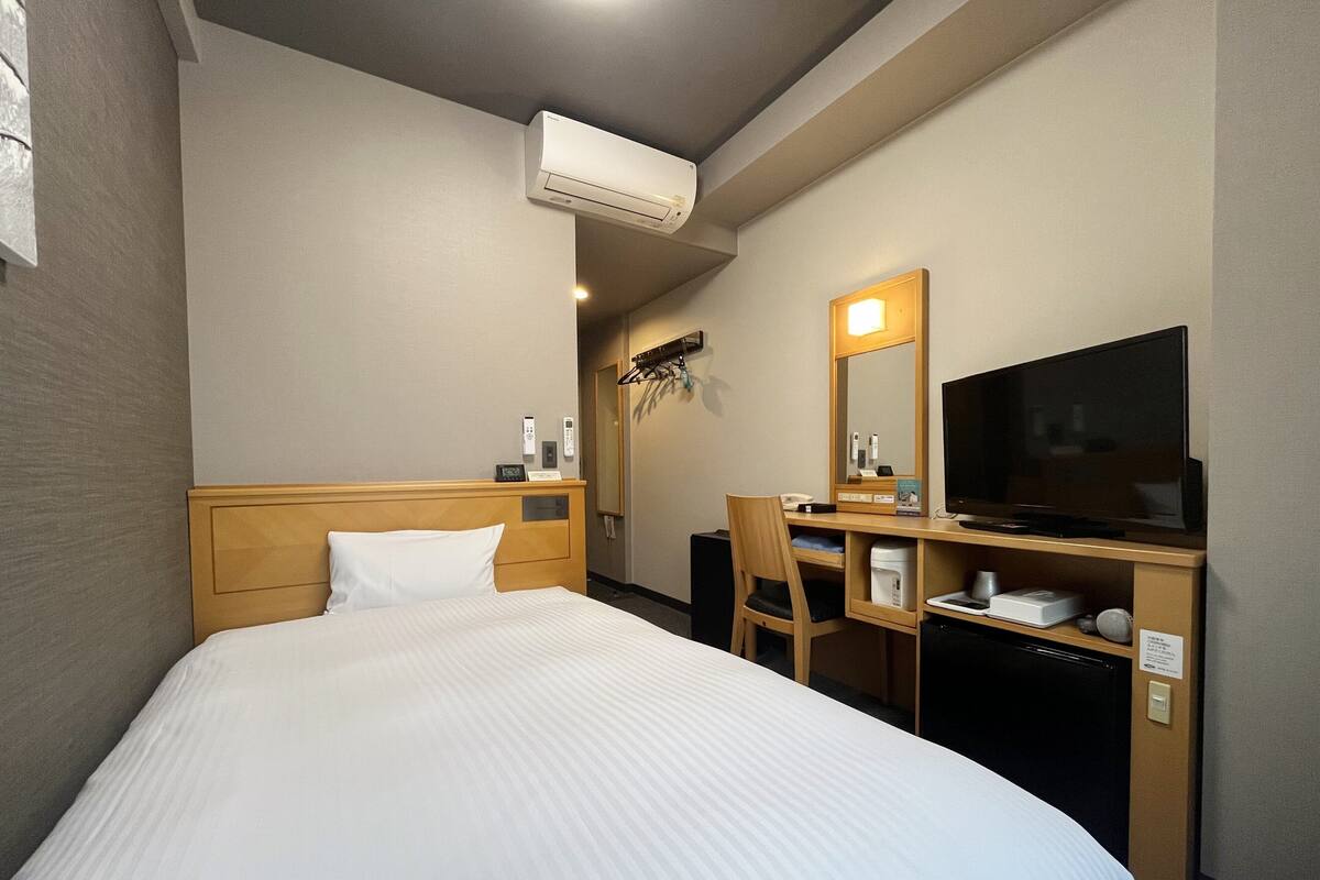 Una habitación de hotel con cama, escritorio con televisor, una silla y un equipo de aire acondicionado.