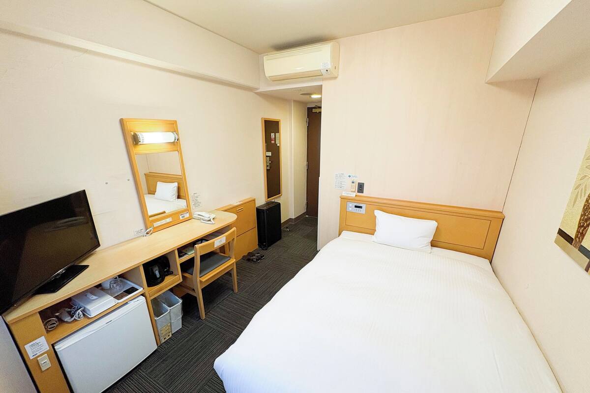 A hotel room with a bed, a desk with a chair, a television, a mini-fridge, a mirror, and an air conditioning unit.
