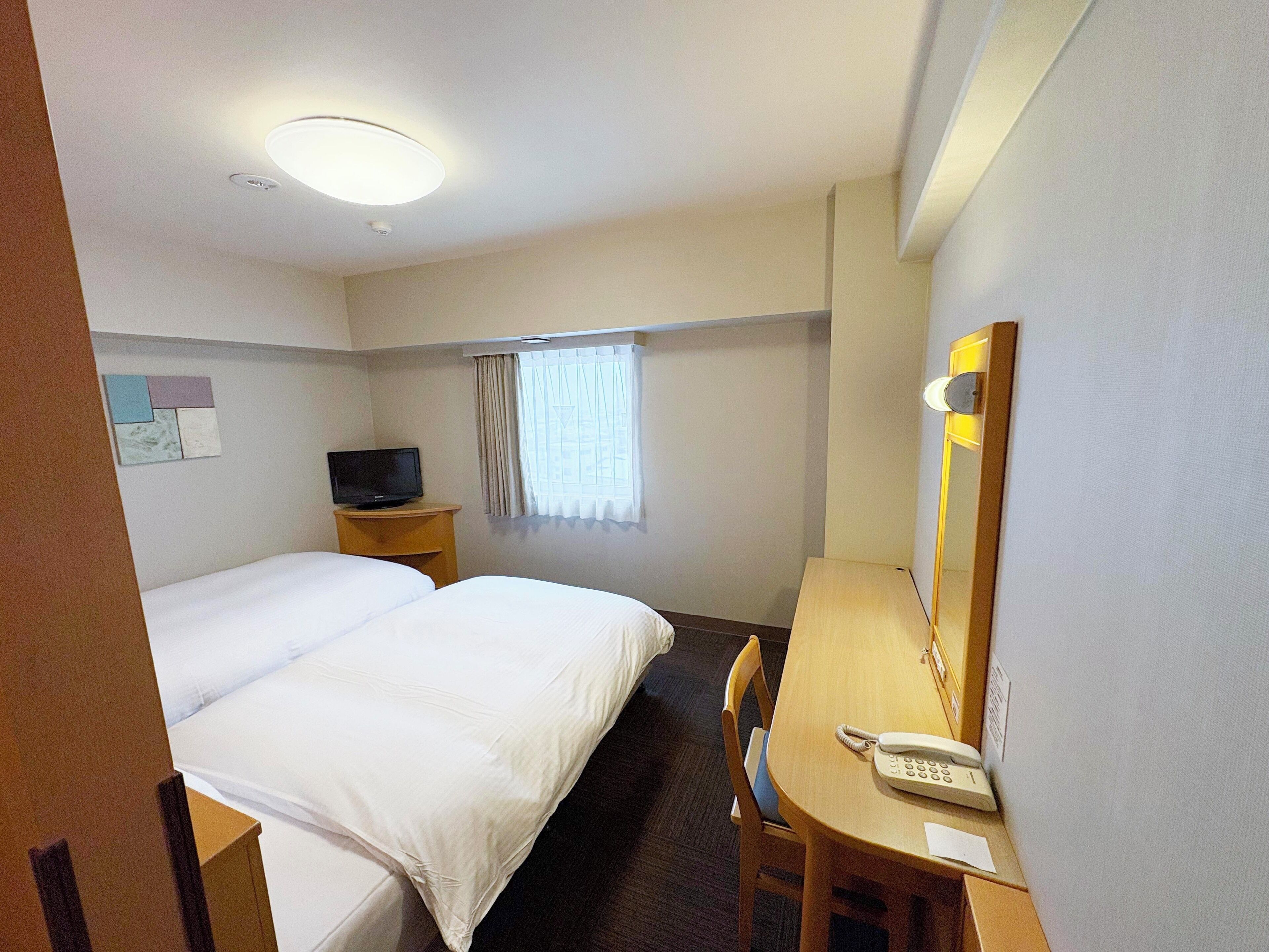 A hotel room with a bed, a desk with a telephone, a television on a side table, and a window with curtains.