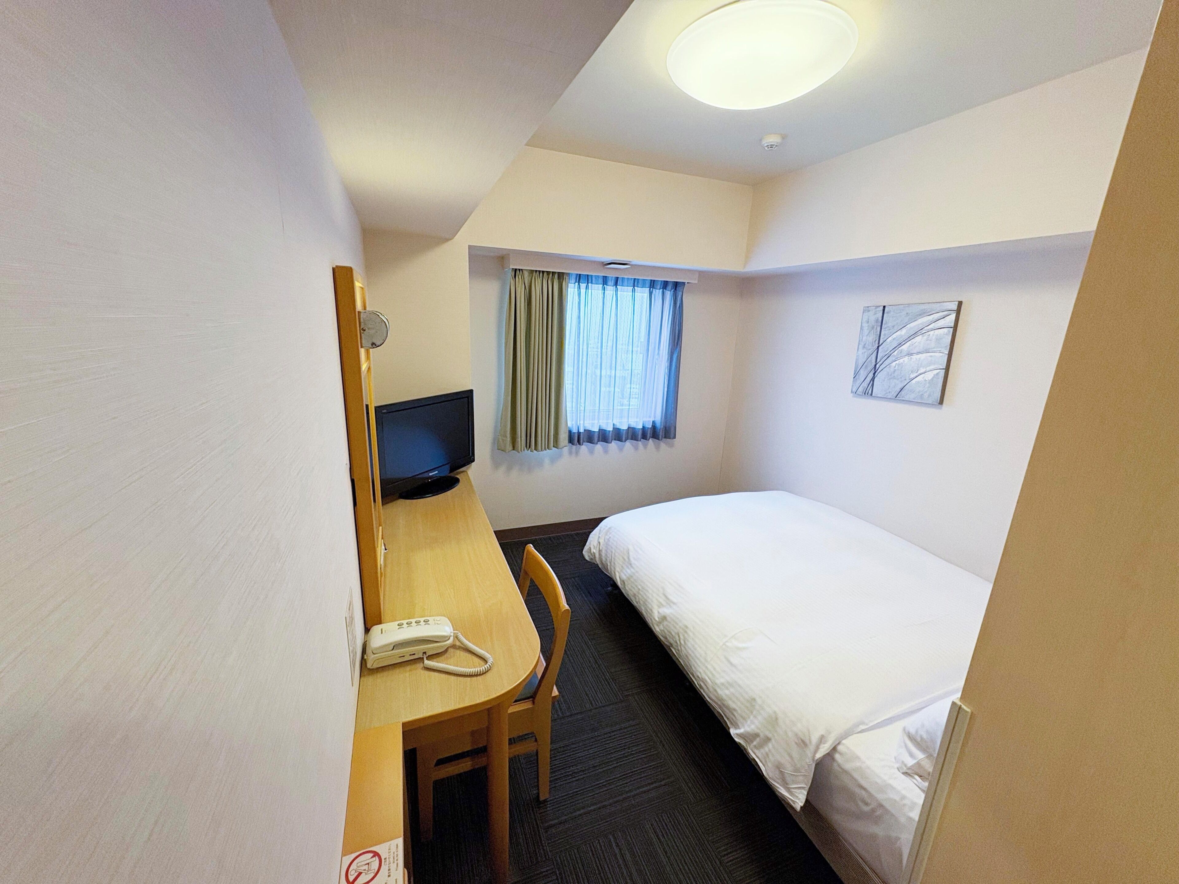 Habitación de hotel con una cama individual, un escritorio con computadora, un teléfono y una ventana con cortinas.