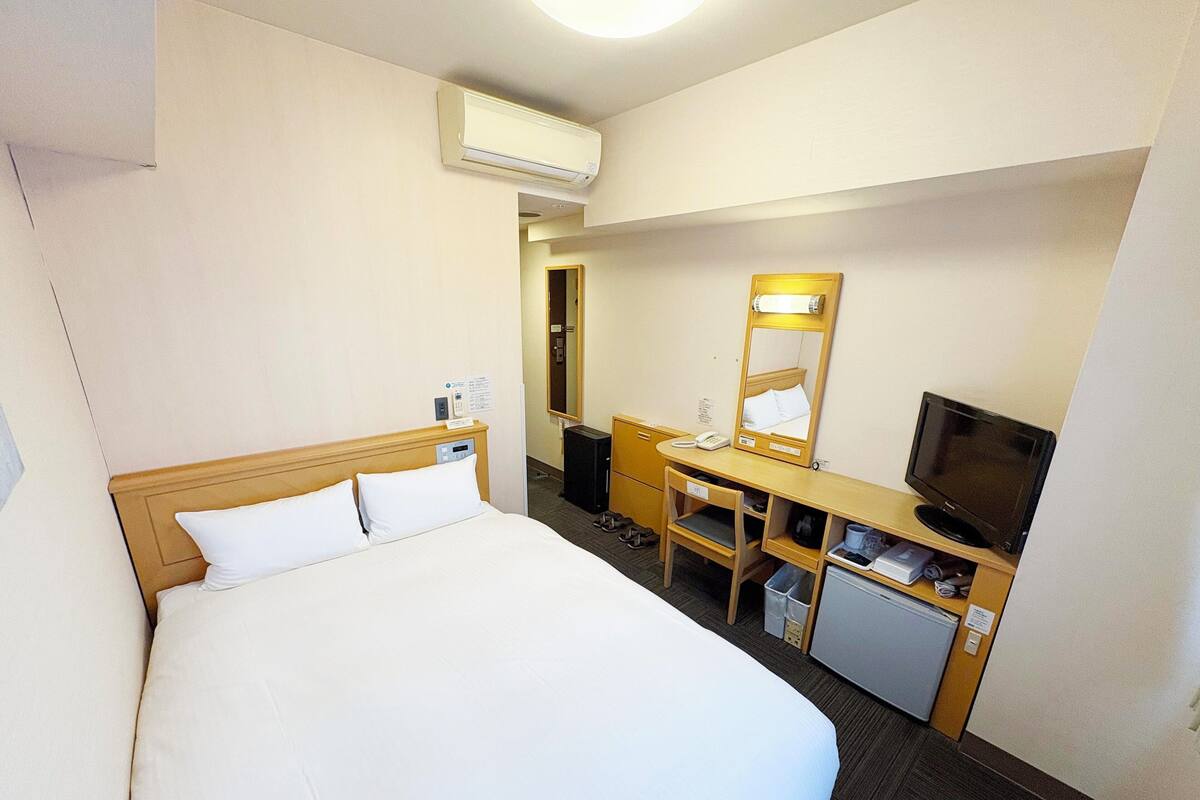 A hotel room with a bed, desk, chair, television, and an air conditioning unit.